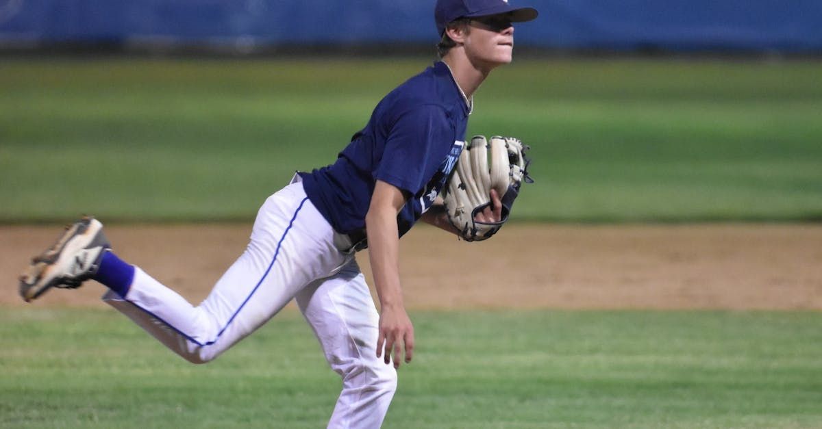 A baseball pitcher in a blue uniform throws a ball on a baseball field; the pitcher is light-skinned and wearing a cap.