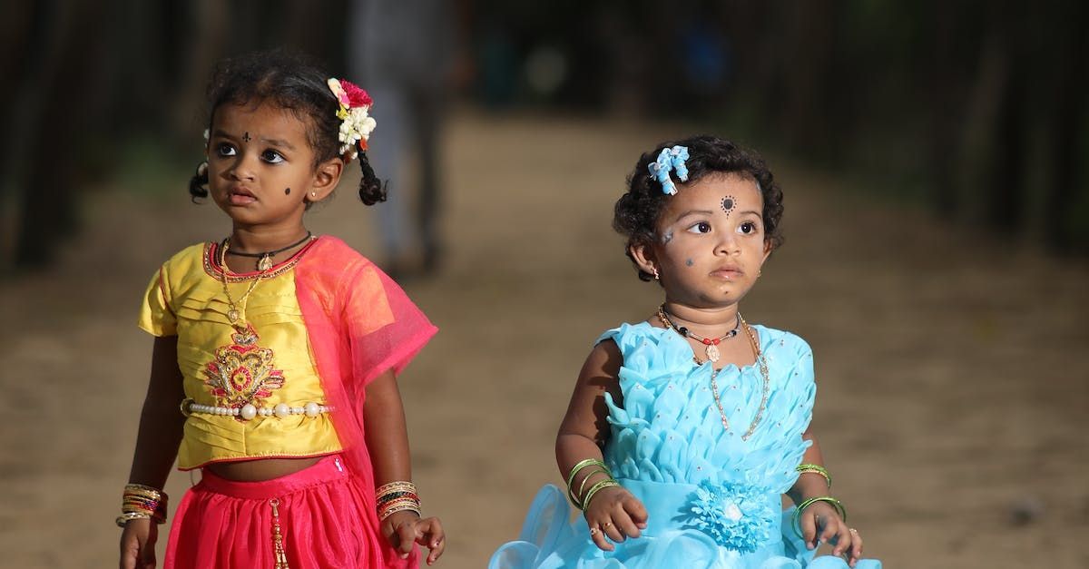 Twee kinderen in kleurrijke jurken staan binnen, de ene in geel en roze, de andere in blauw.