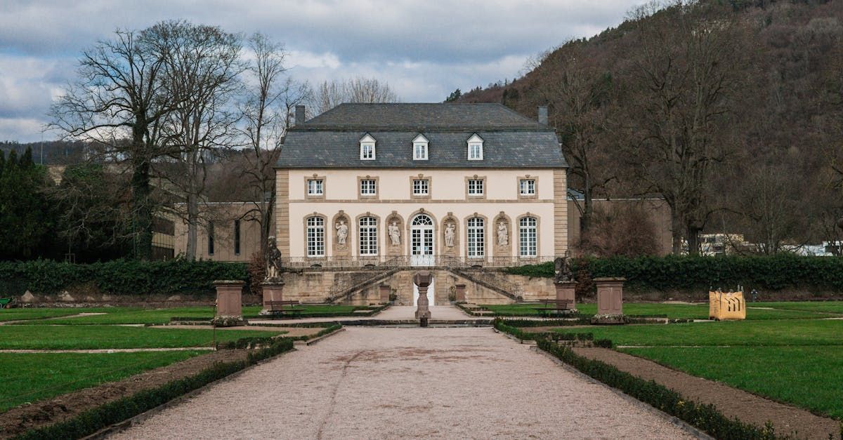 Un grand manoir de couleur claire avec un jardin formel et une colline en arrière-plan.