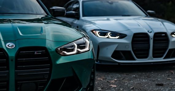 A green and a white bmw m3 are parked next to each other on a gravel road.