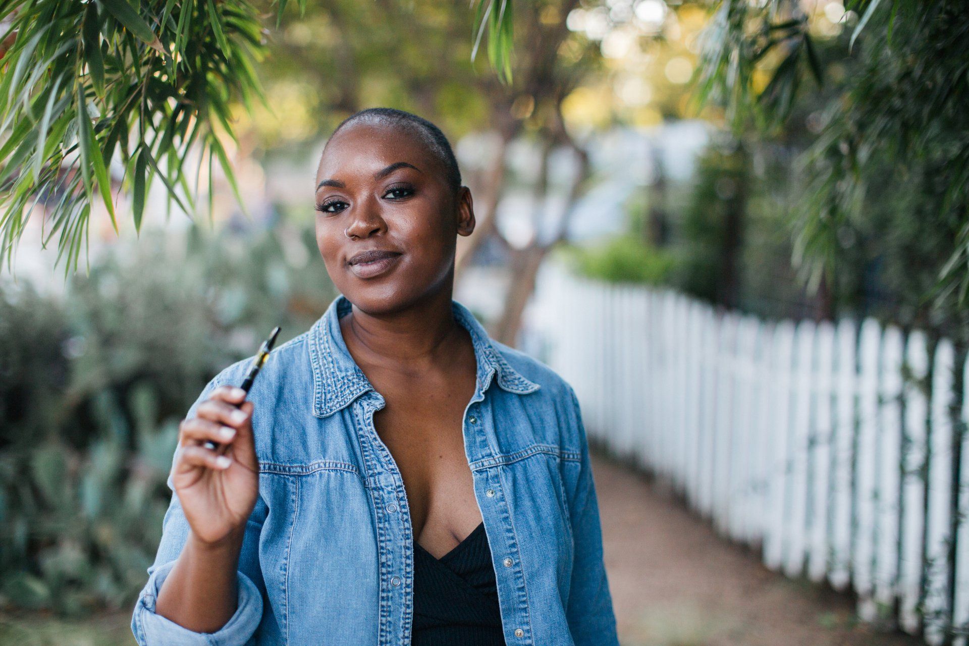 Woman with shaved head, smiling, holding pen-like object, wearing denim jacket, outdoors near white fence.