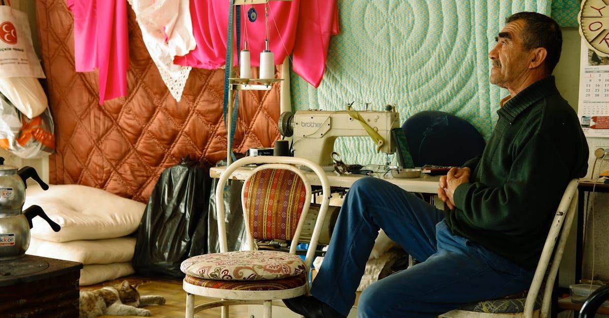 Man in a sewing room, seated, looking at something. Bright clothing hangs. Sewing machine on a table.