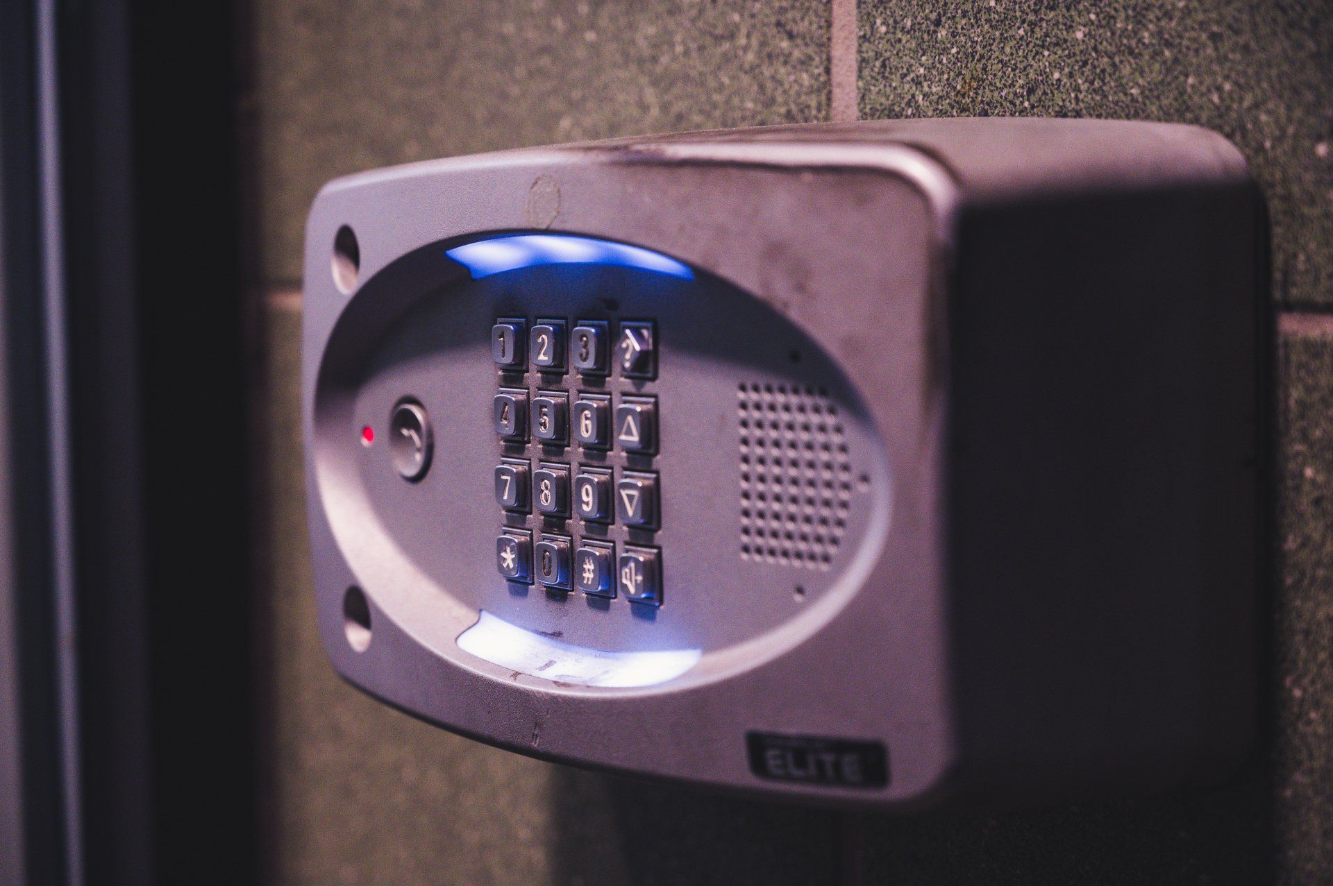 Gray intercom keypad on a green-tiled wall with illuminated blue buttons and a red light.