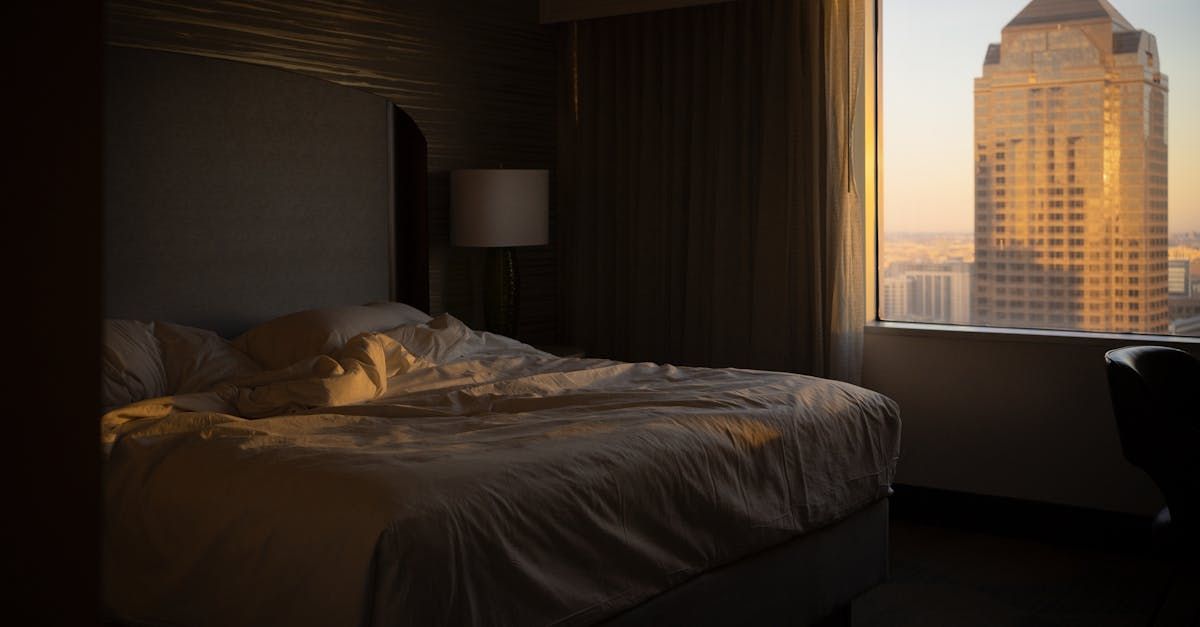 Bedroom with unmade bed, sunlight streams through a window, illuminating a tall building in the distance.