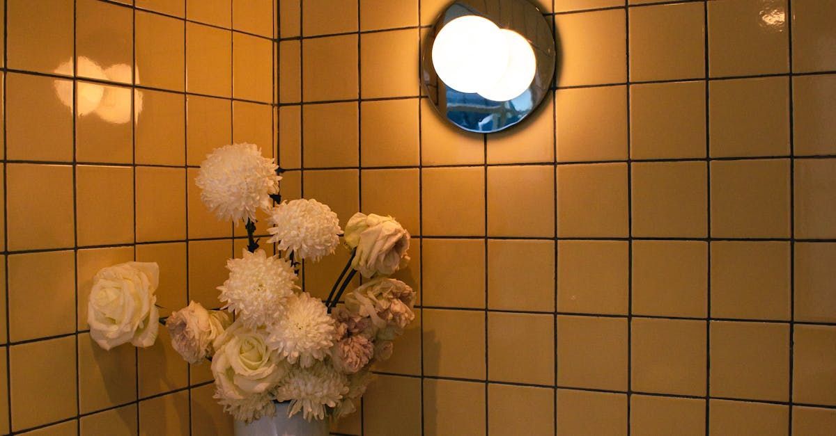 White flowers in a vase against a tan tiled wall with a round light fixture above.