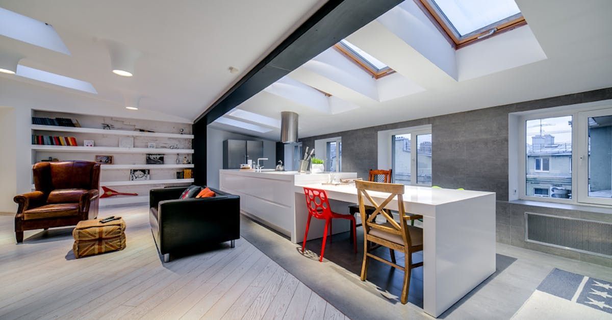 Bright modern kitchen and lounge with skylights, white island, red chairs, and gray sectional sofa.