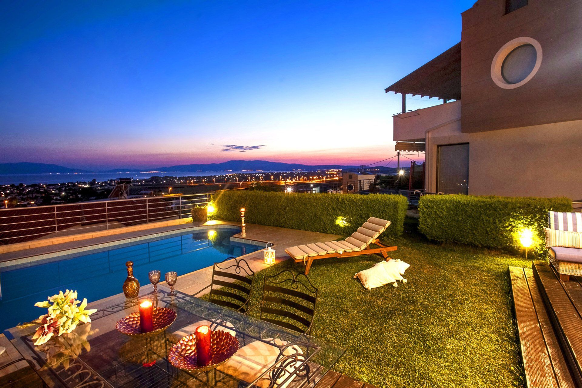 Evening view of a villa's pool and garden, overlooking a city at dusk, with ambient lighting and outdoor furniture.