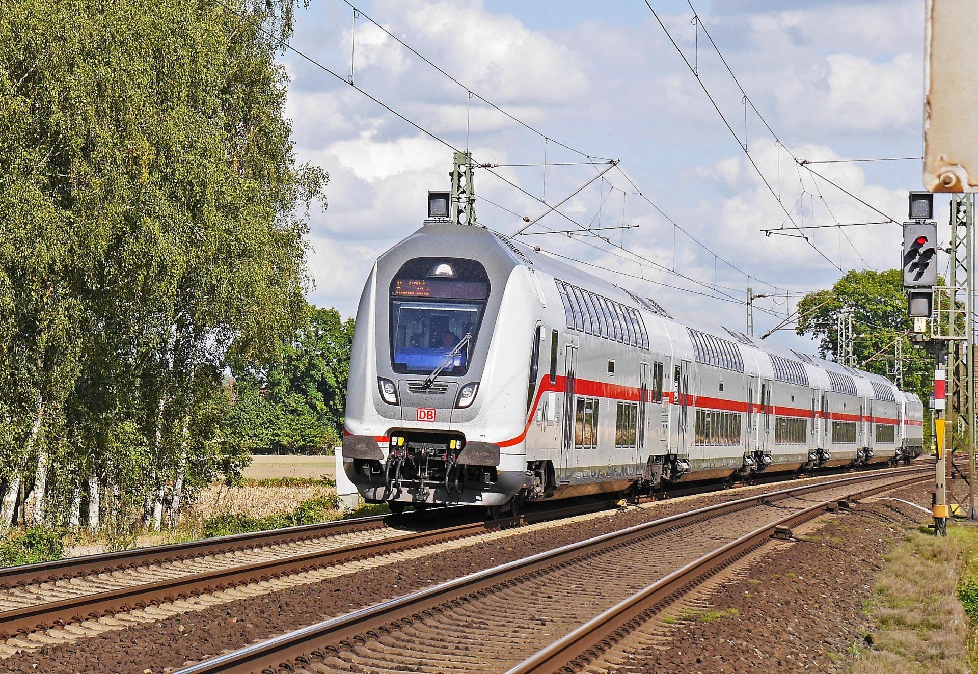 A modern silver and red passenger train traveling on tracks, passing trees and signals.