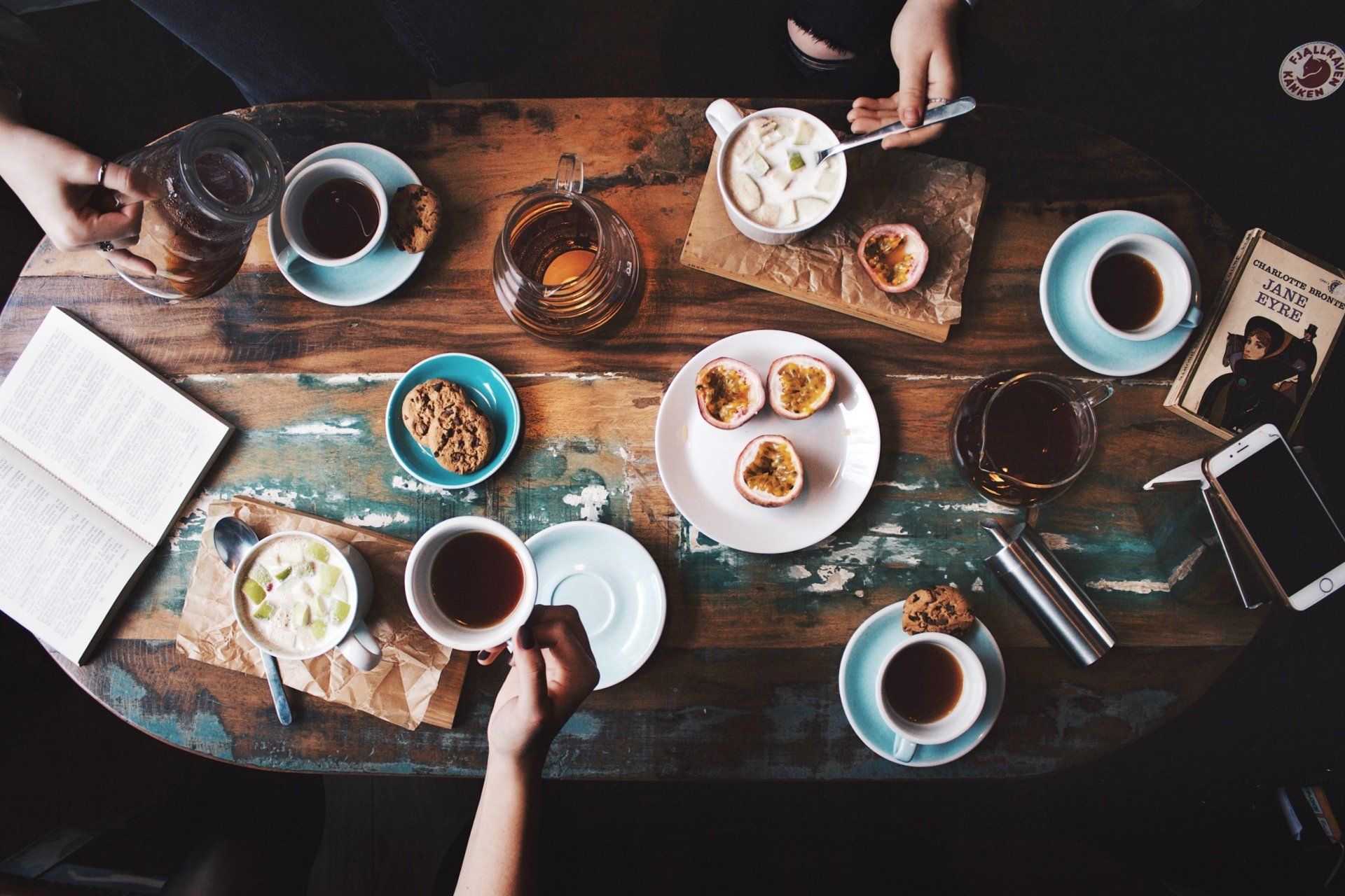 Een opname van bovenaf van handen rond een rustieke houten tafel, gedekt met thee, koffie, kleine hapjes, een boek en een smartphone.