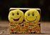 Two yellow smiley face plush toys in a box decorated with emoji faces, outdoors.