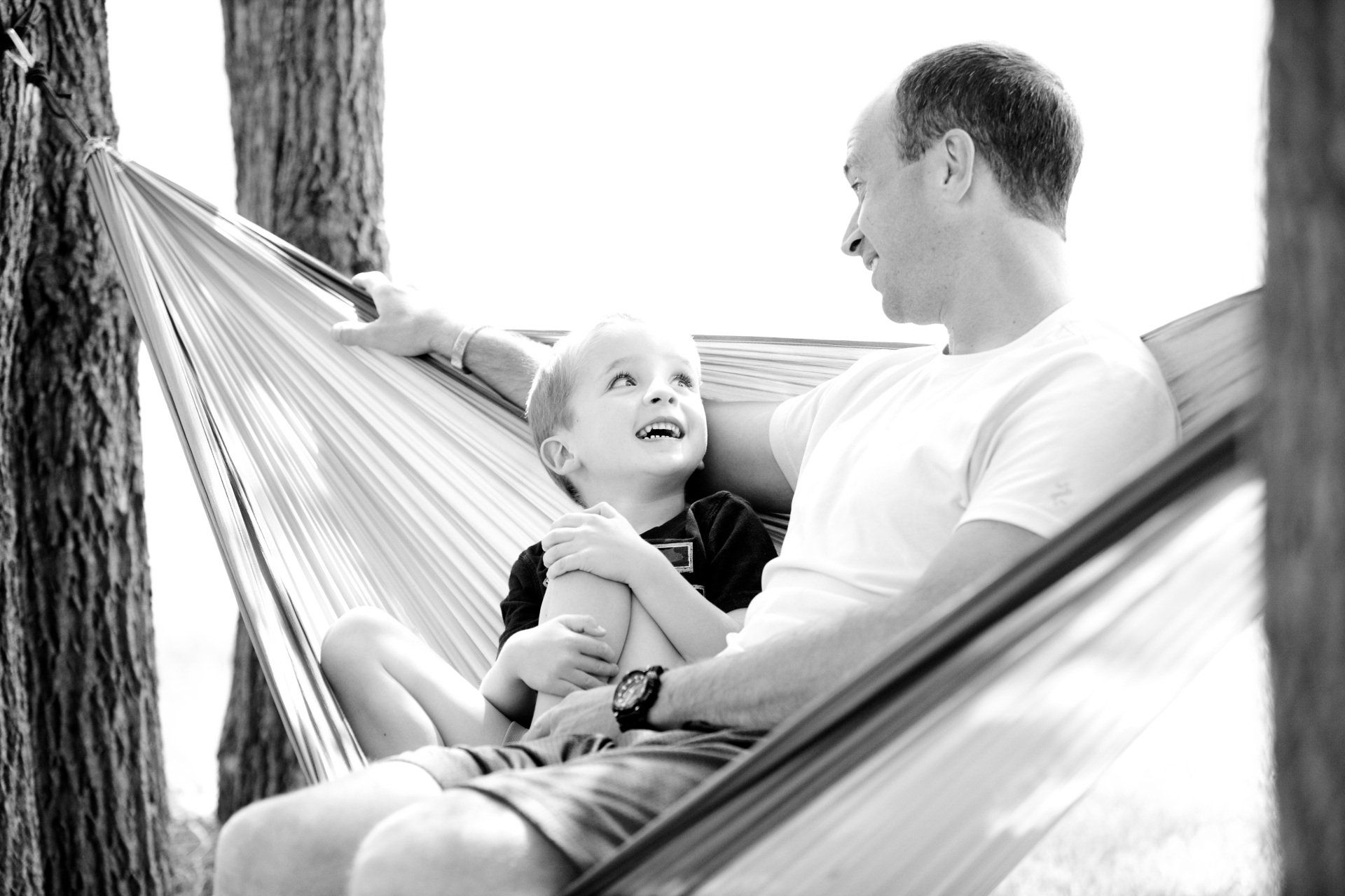 A parent and child laughing and looking at each other while relaxing together in a fabric hammock between two trees.