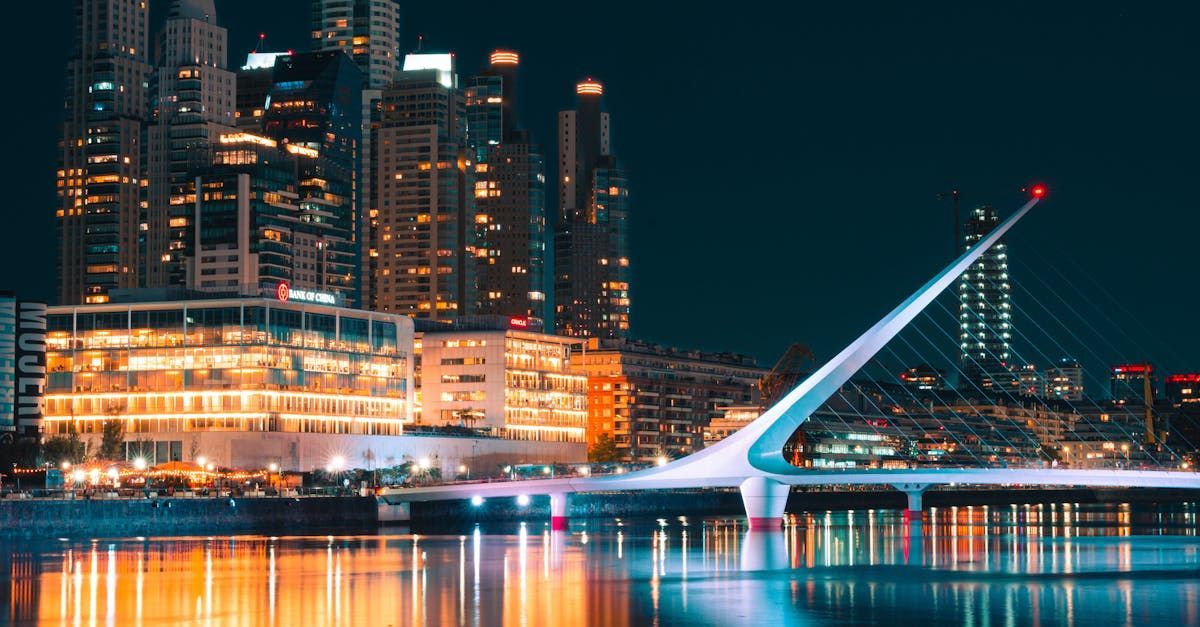 Night view of illuminated Puente de la Mujer pedestrian bridge in Buenos Aires, Argentina, with city skyline.
