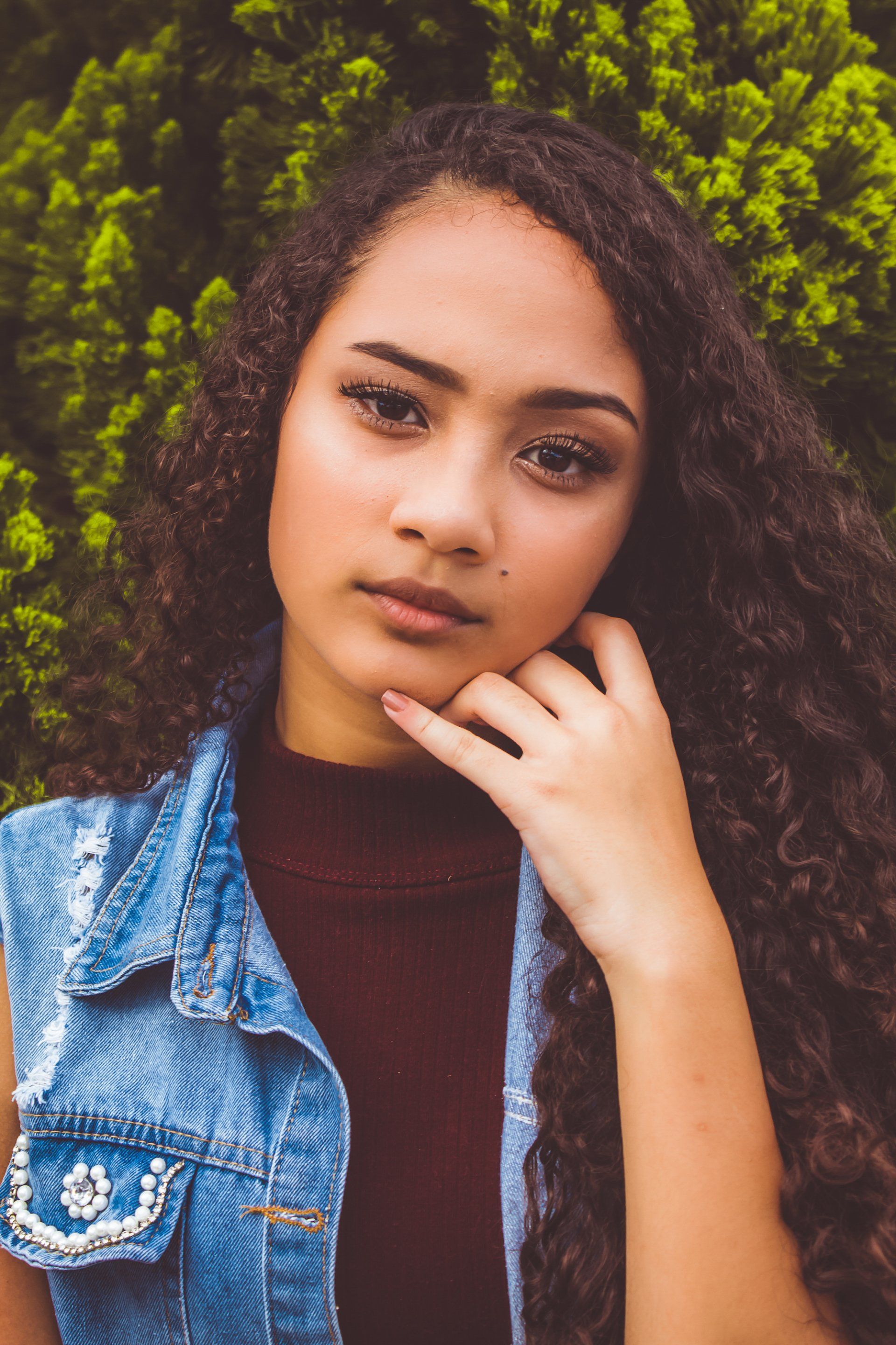 A young woman with long curly hair is wearing a denim vest and a turtleneck.