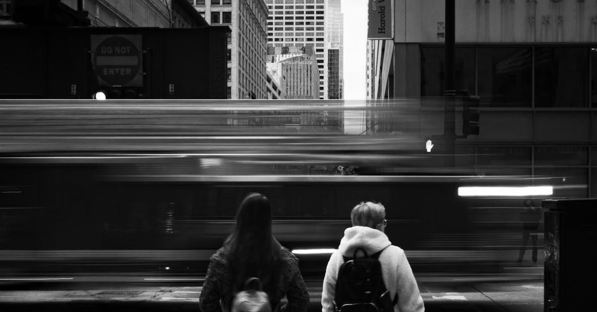 Two people with backpacks stand on a city platform as a train blurs past in black and white.