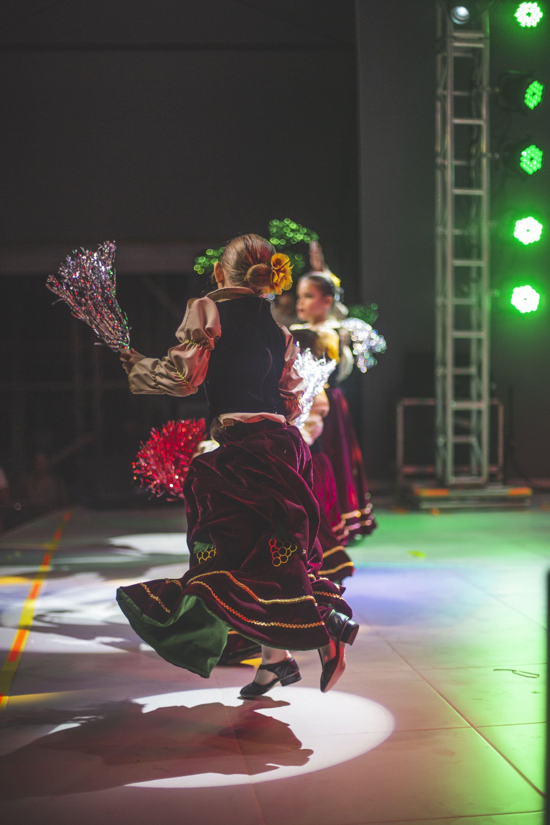 Dancers on a lit stage perform in traditional flowing, dark velvet skirts and patterned tops, holding decorative props.