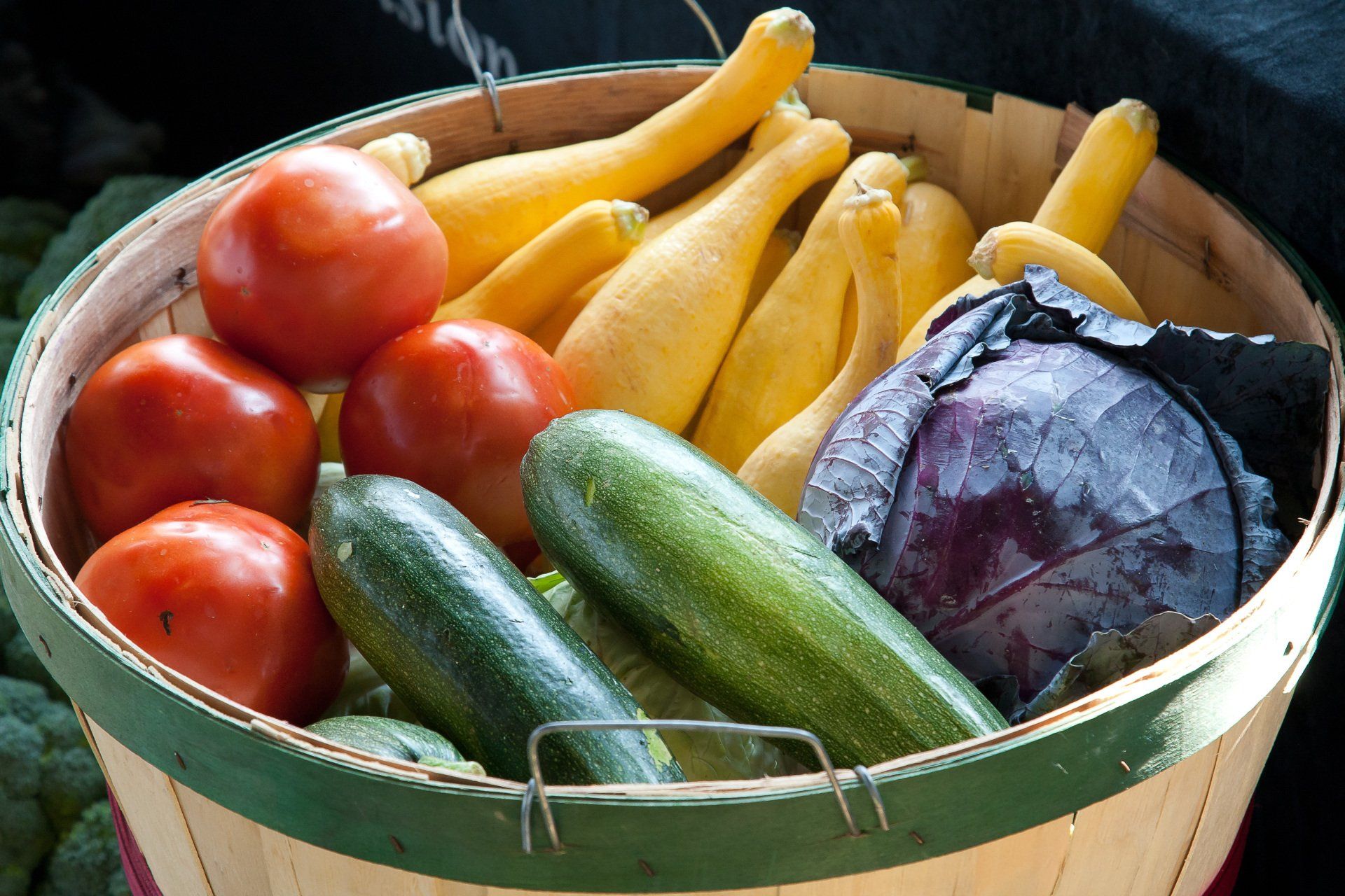 Basket of assorted fresh vegetables, including tomatoes, cucumbers, yellow squash, and a purple cabbage
