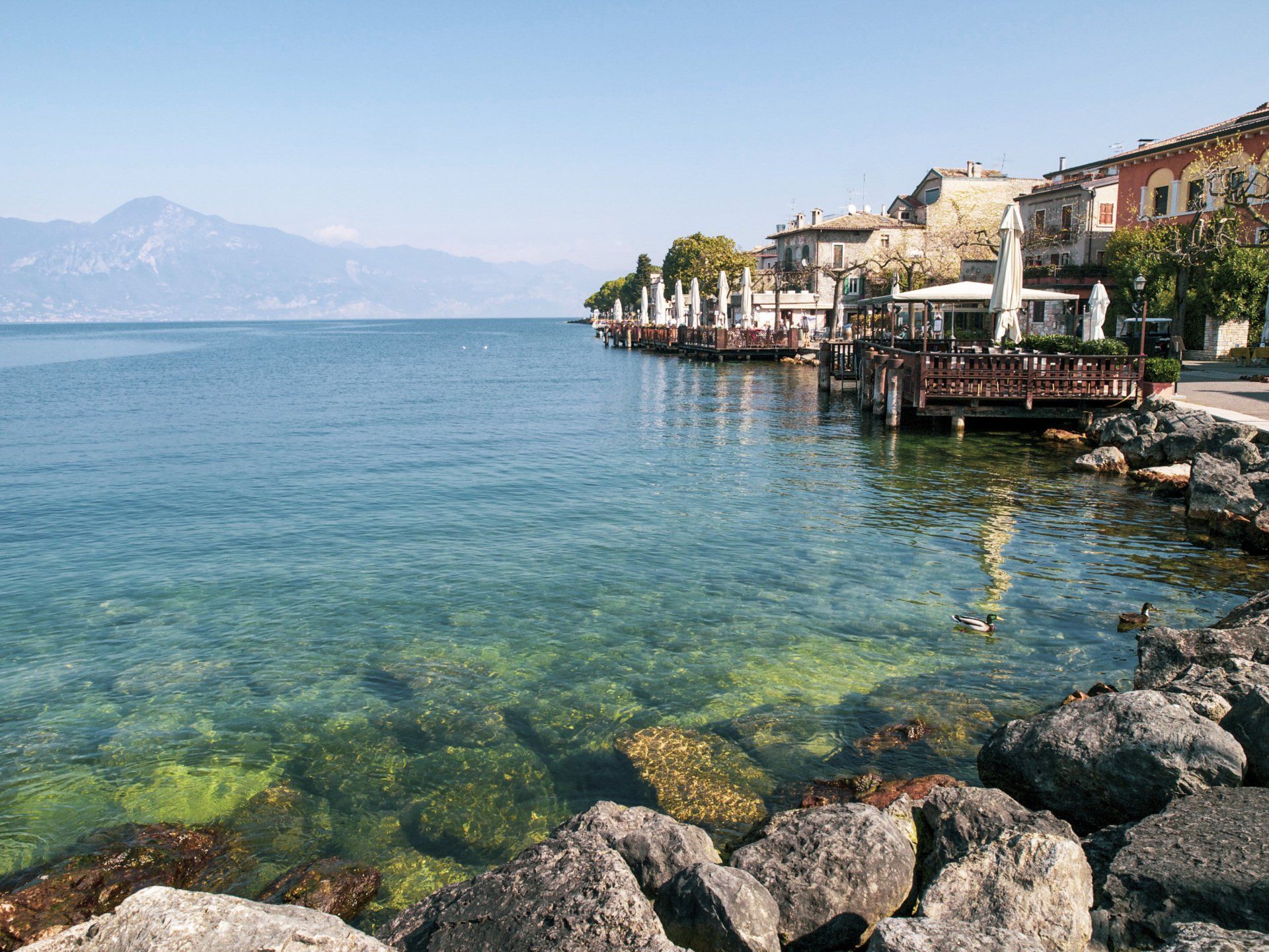 Rocky shoreline with clear turquoise water, buildings, and mountains in the background.