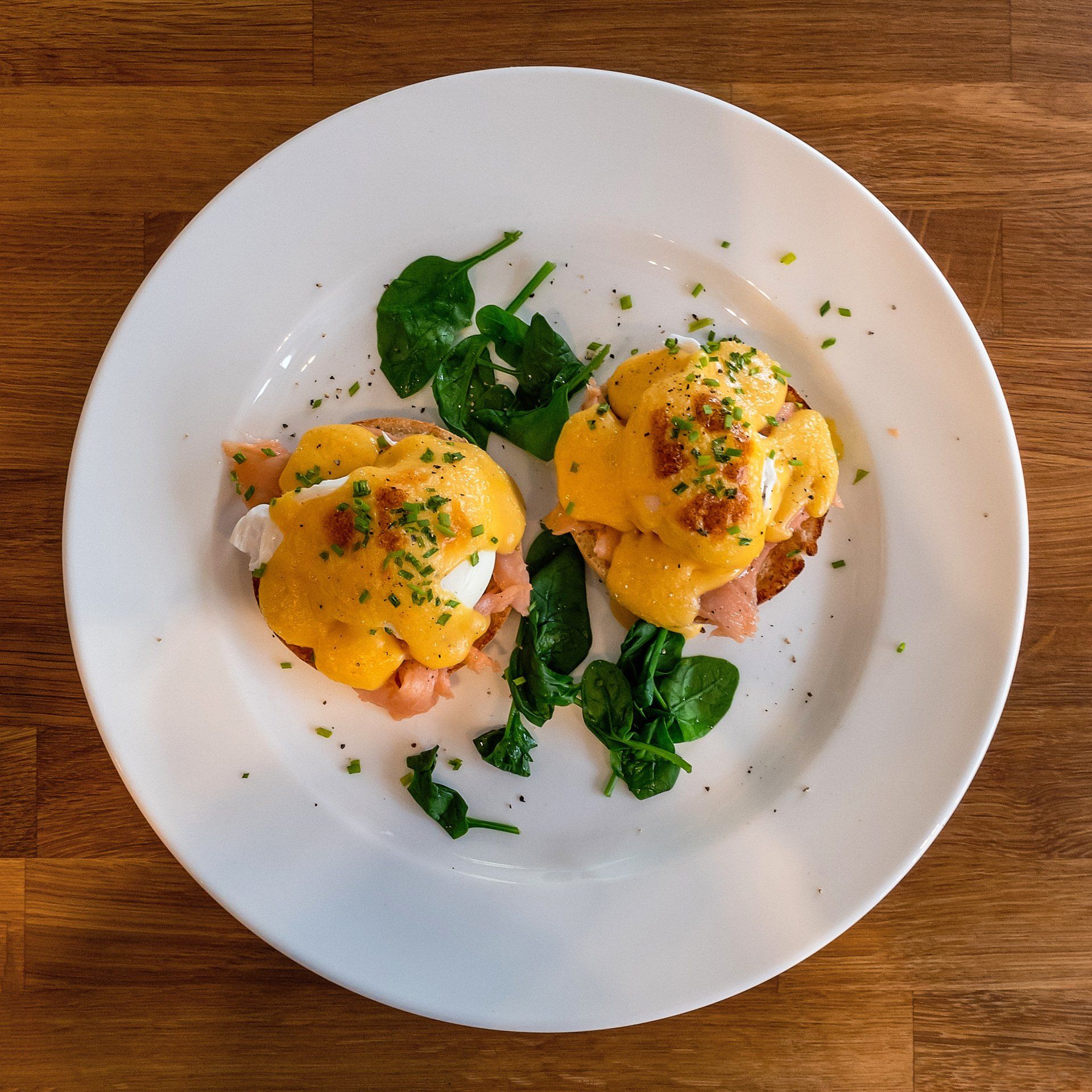 Two Eggs Benedict with smoked salmon and a side of spinach, topped with hollandaise sauce on a white plate.