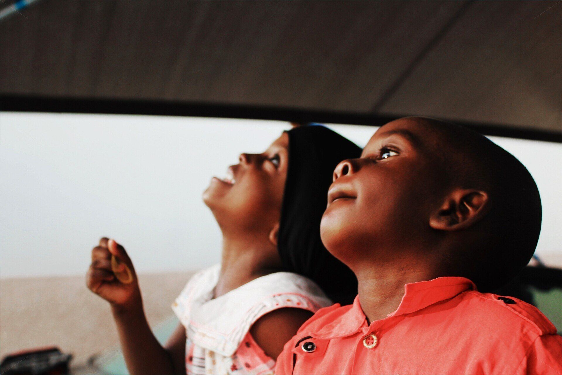 Two children looking upwards with hopeful expressions; one in a pink shirt, the other in a white dress.