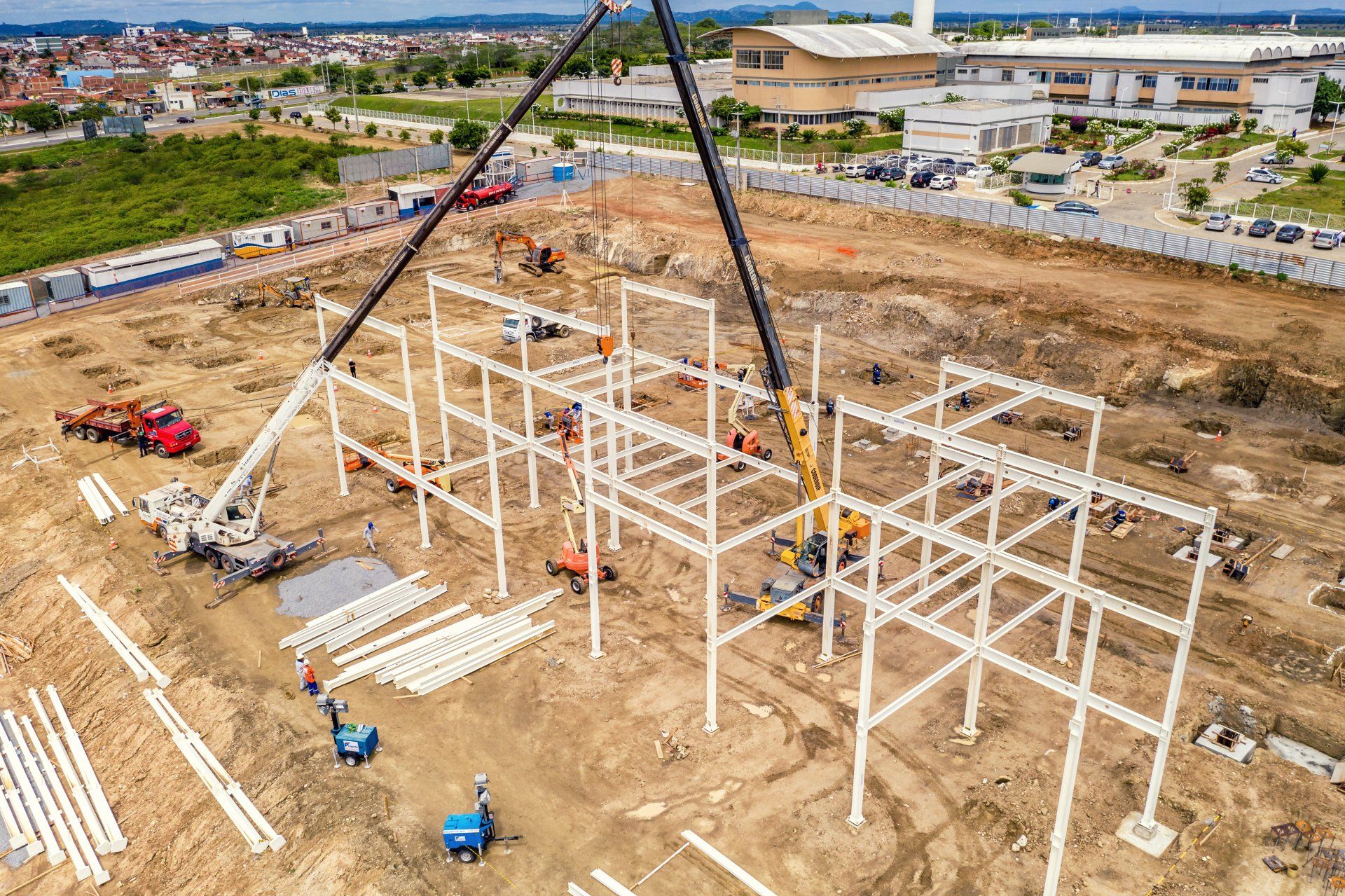 Construction site with cranes lifting steel beams to erect a building frame; workers in action.