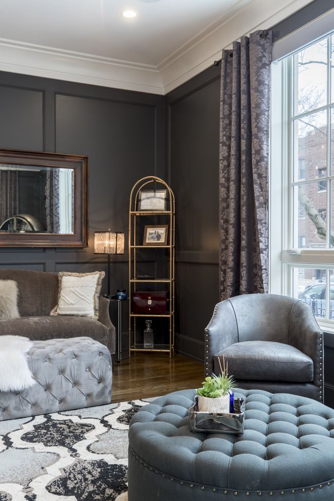 Elegant bedroom corner with gray walls, tufted ottoman, armchair, bed, mirror, and tall window with curtains