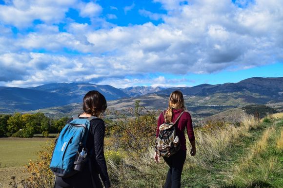Two people hiking on a path overlooking mountains under a blue, cloudy sky.