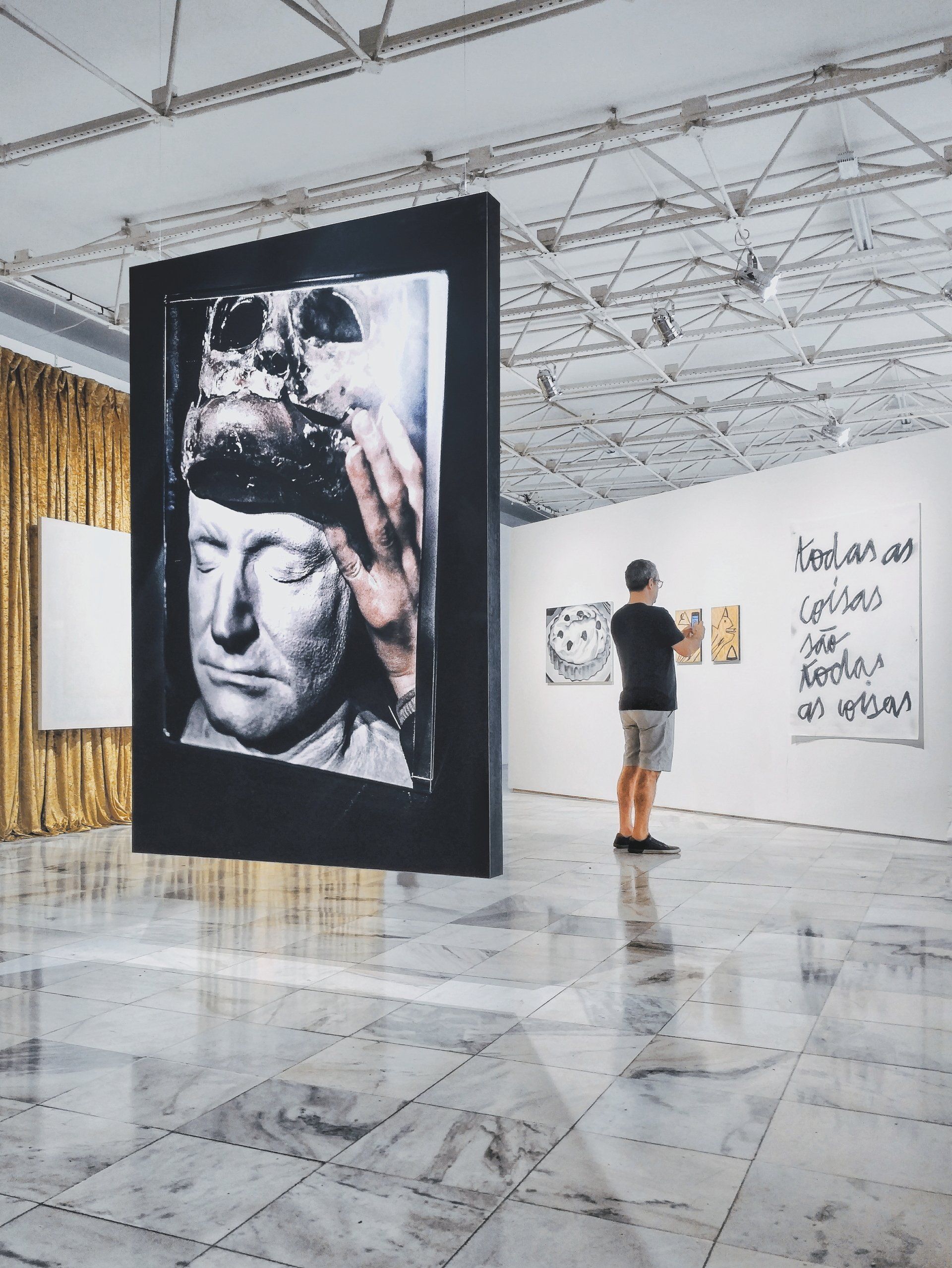 Art gallery: large black-framed artwork suspended; viewer examines other pieces on white wall.