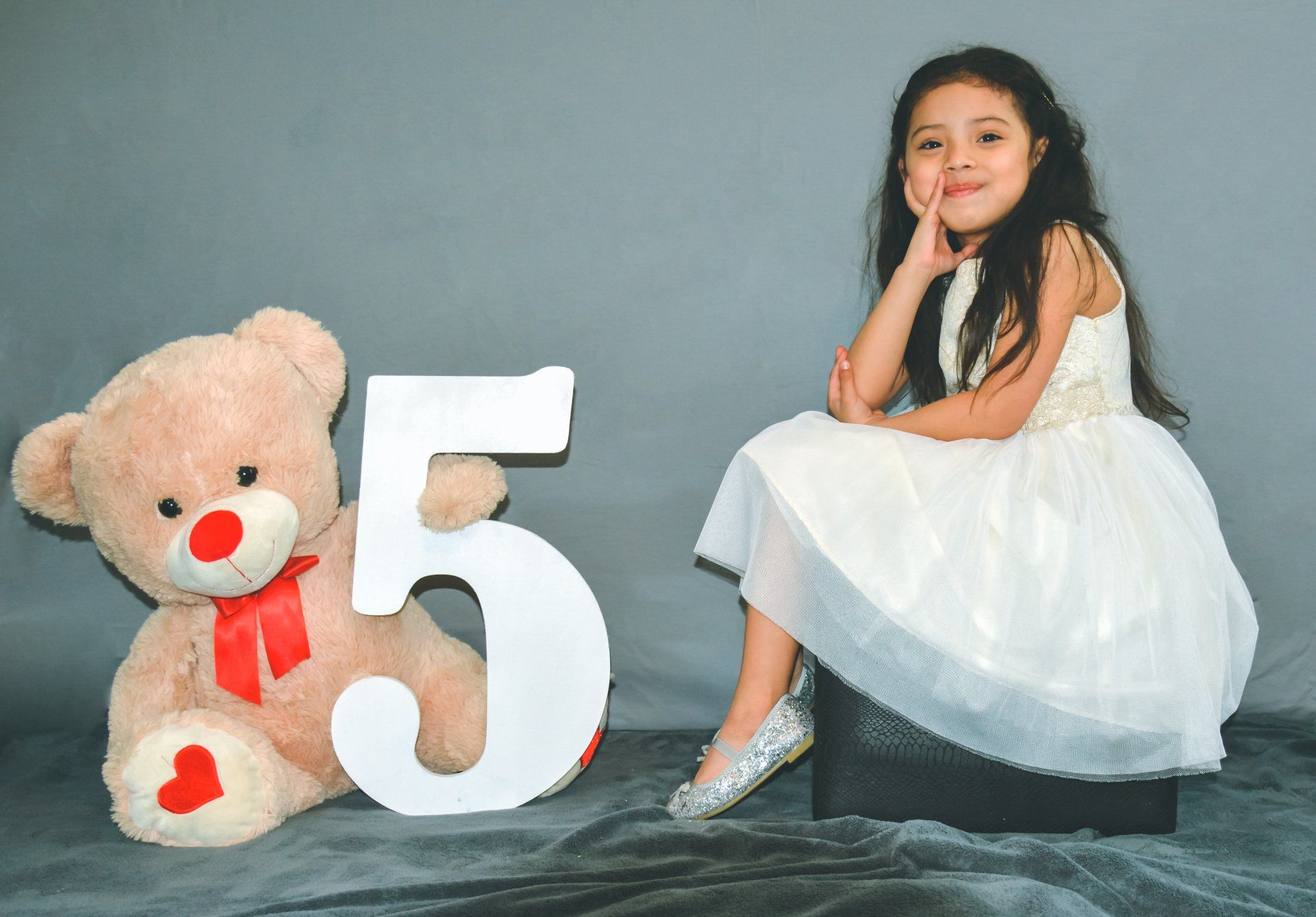 A young child in a white dress sits next to a large teddy bear and a white number five on a gray background.