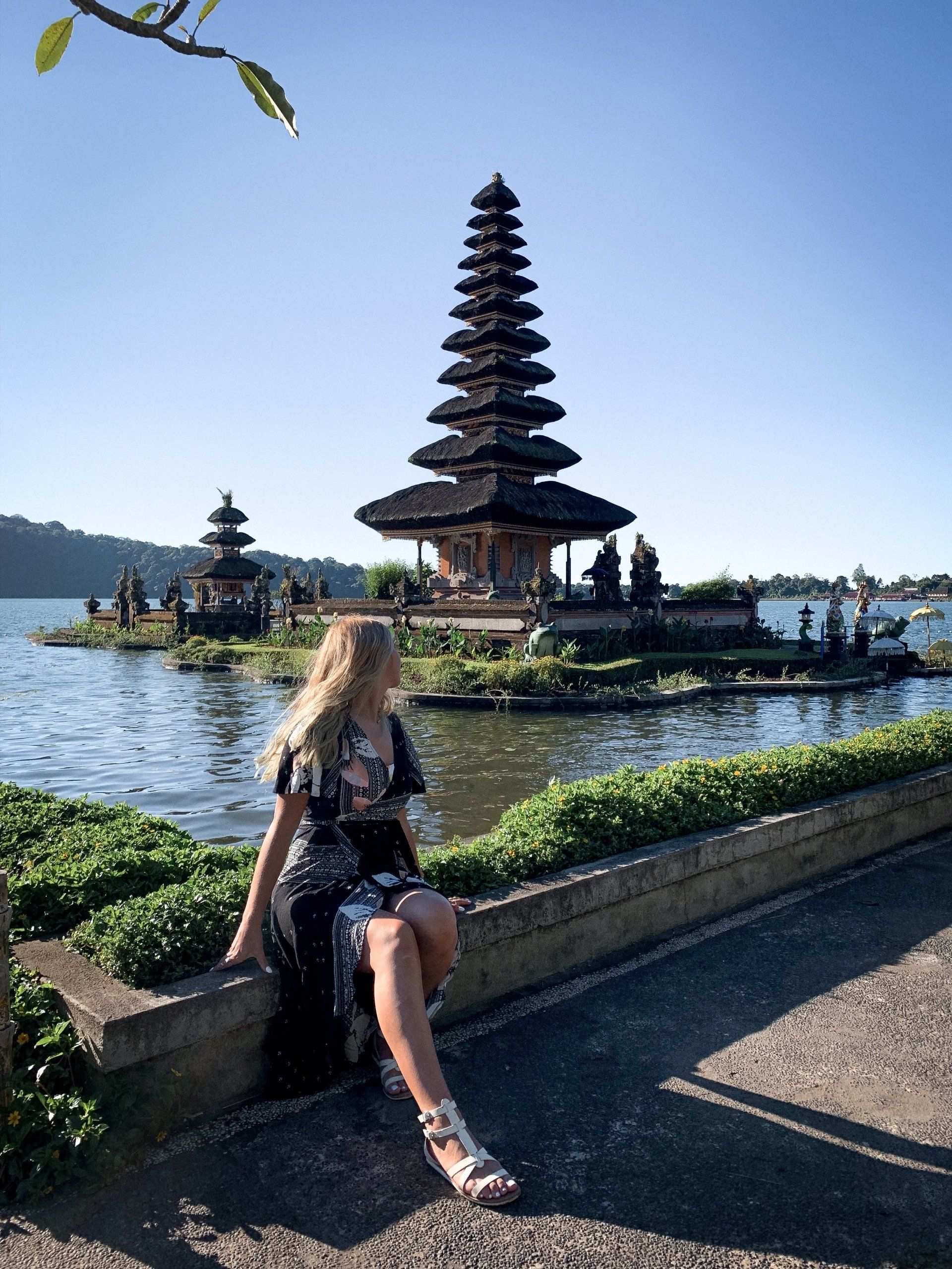 A woman is sitting on a wall next to a lake with a temple in the background.