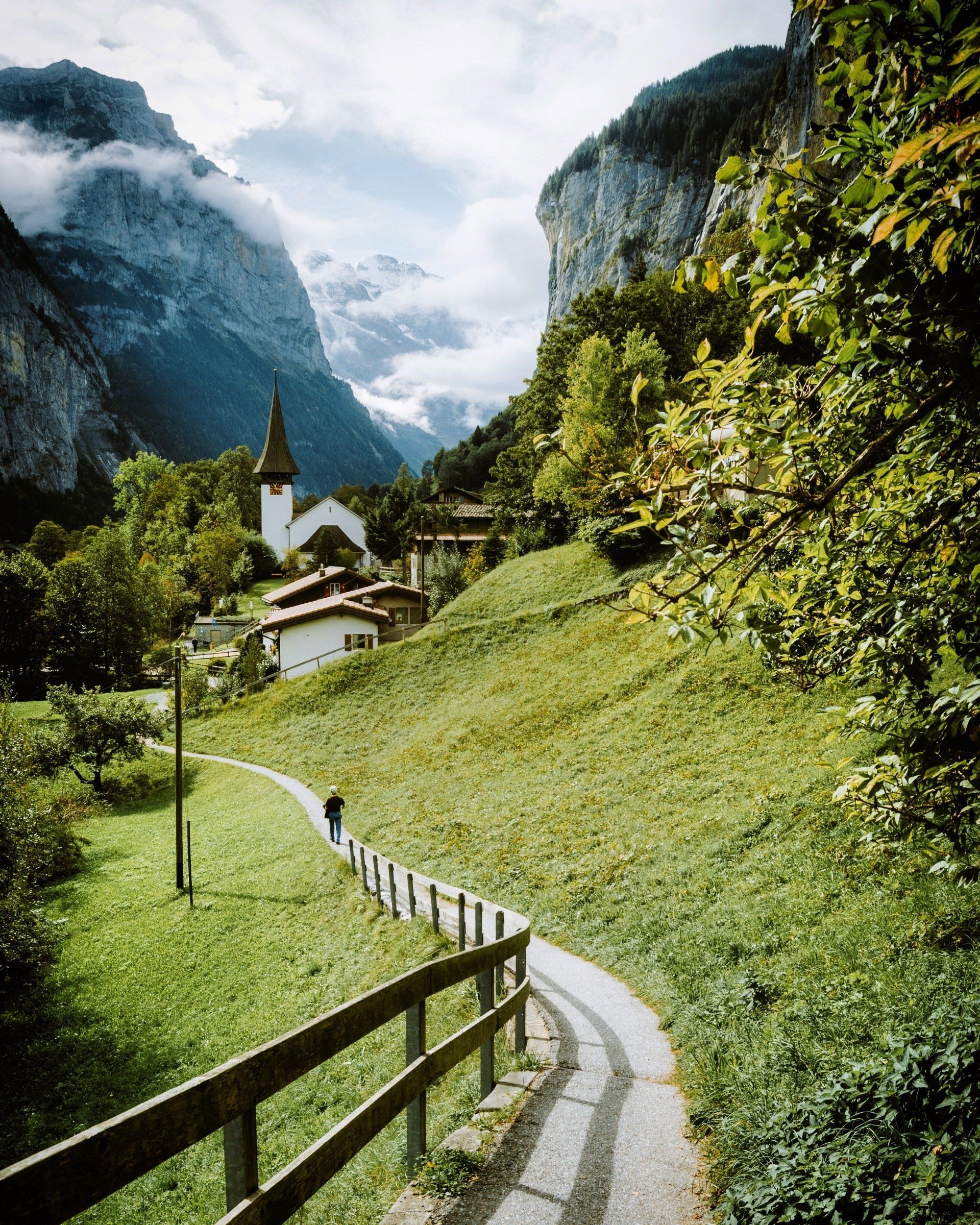 A person walks along a winding path toward a church in a lush, mountainous Swiss valley under a cloudy sky.