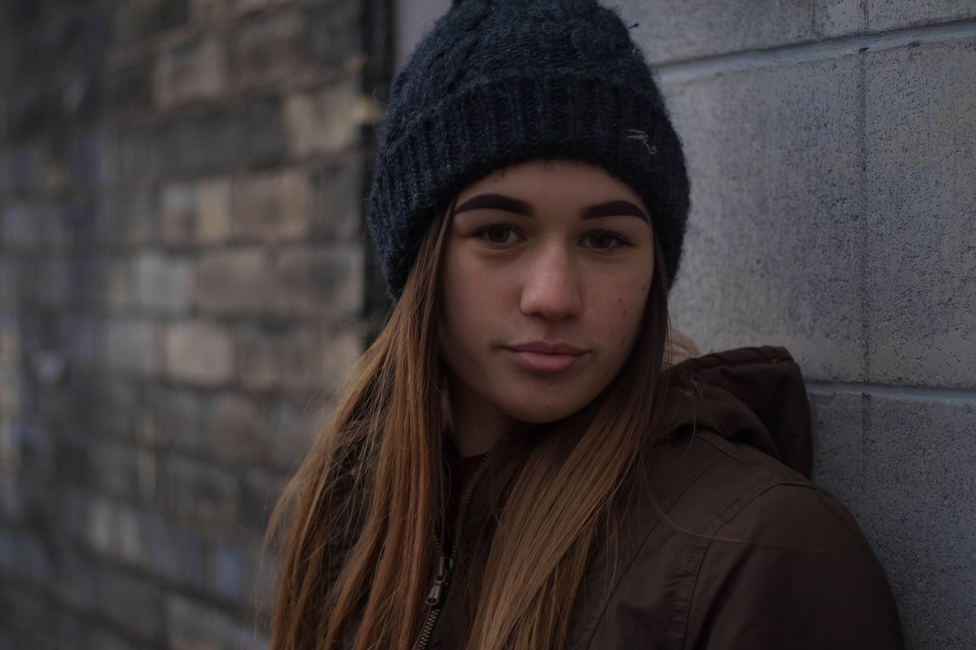 A young woman wearing a beanie and a jacket is leaning against a brick wall.