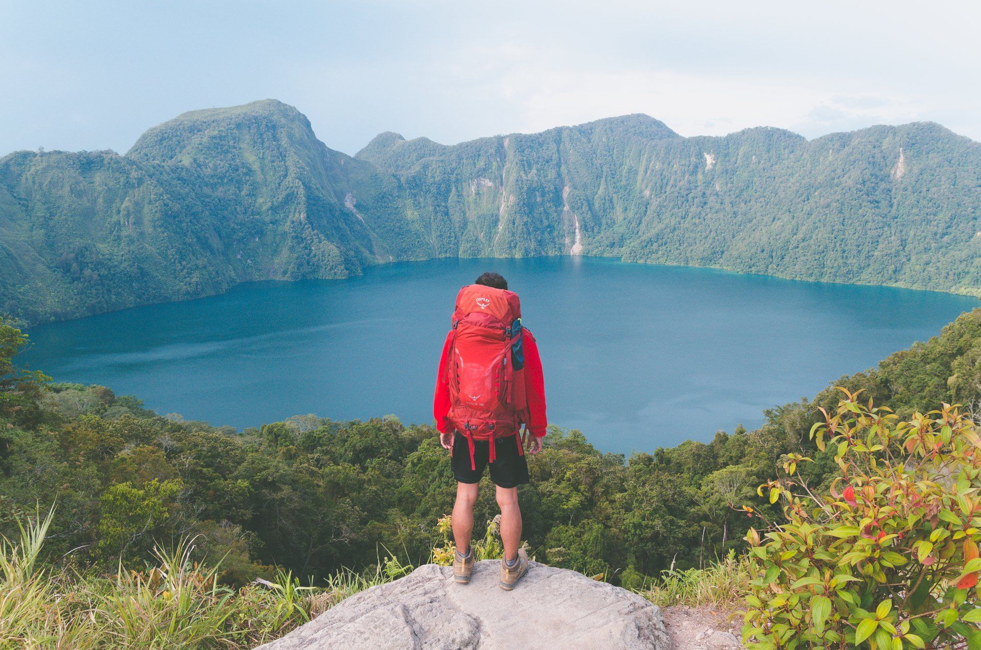 A hiker with a large red backpack stands on a rock overlook, gazing at a vast, circular crater lake surrounded by mountains.