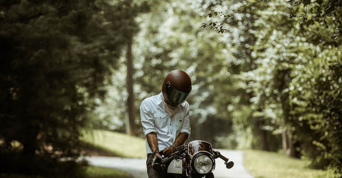 Eine Person mit braunem Helm und weißem Hemd steht auf einem Motorrad an einer von Bäumen gesäumten Straße.