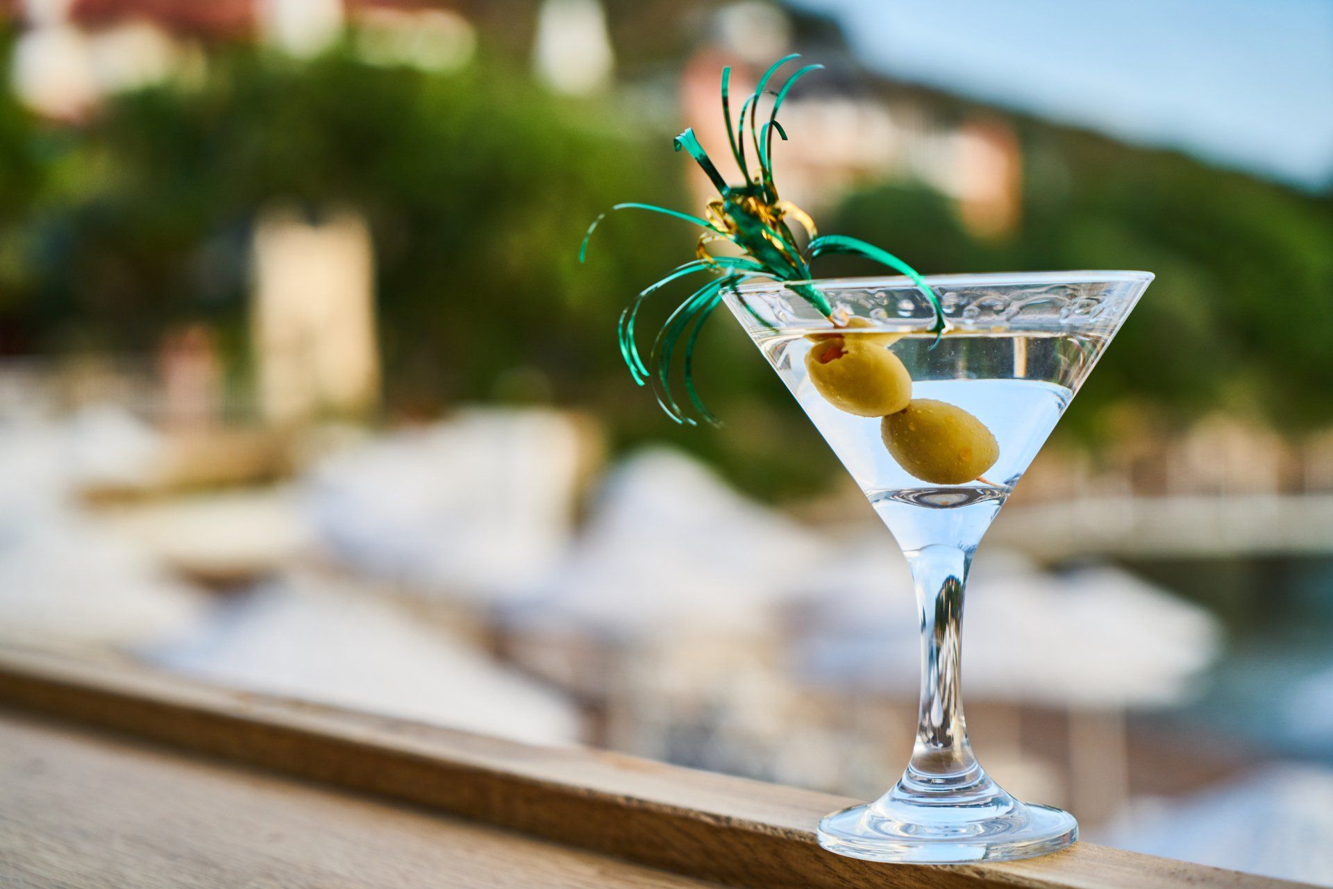 Martini glass with olives, garnished, on a wooden railing, blurry background of buildings and greenery.