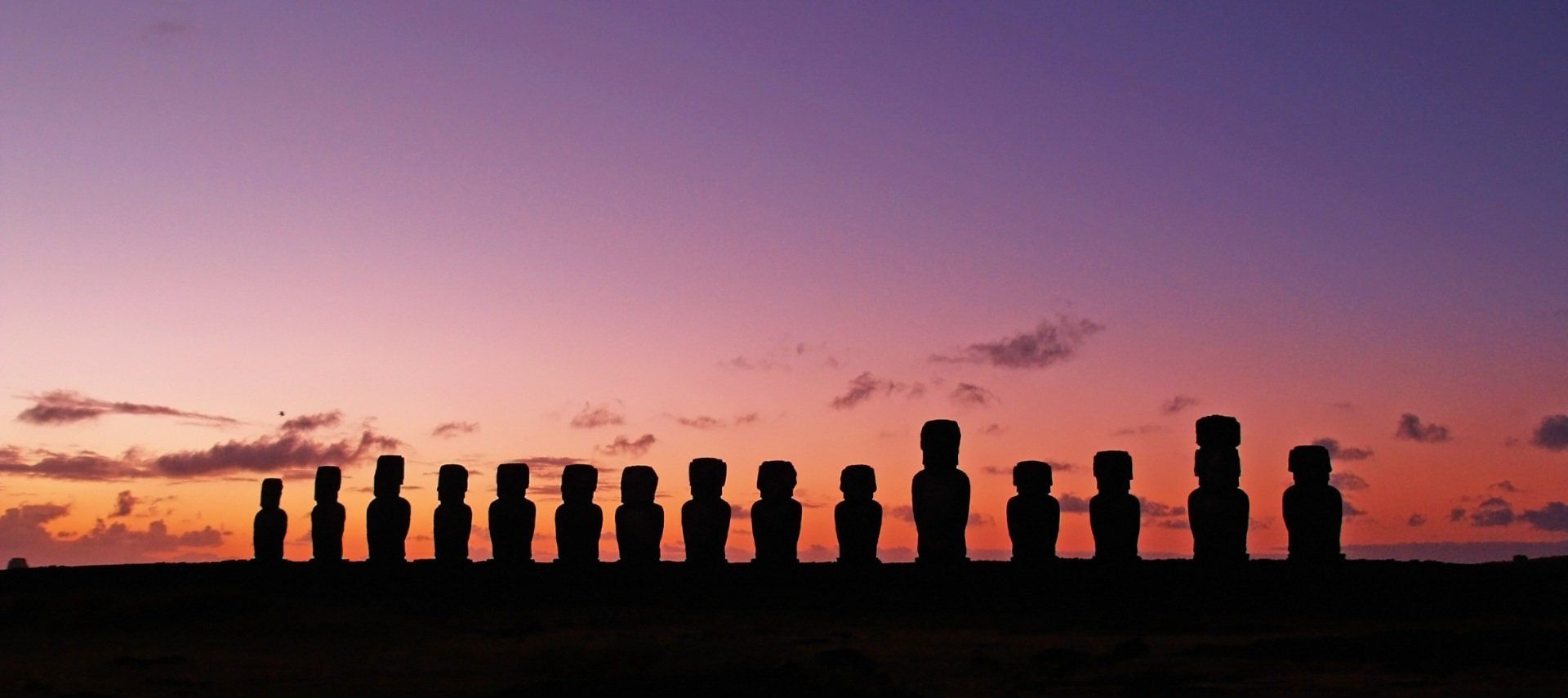 Silhuetter af moai-statuer på en græsklædt bakke mod en levende solnedgangshimmel på Påskeøen.