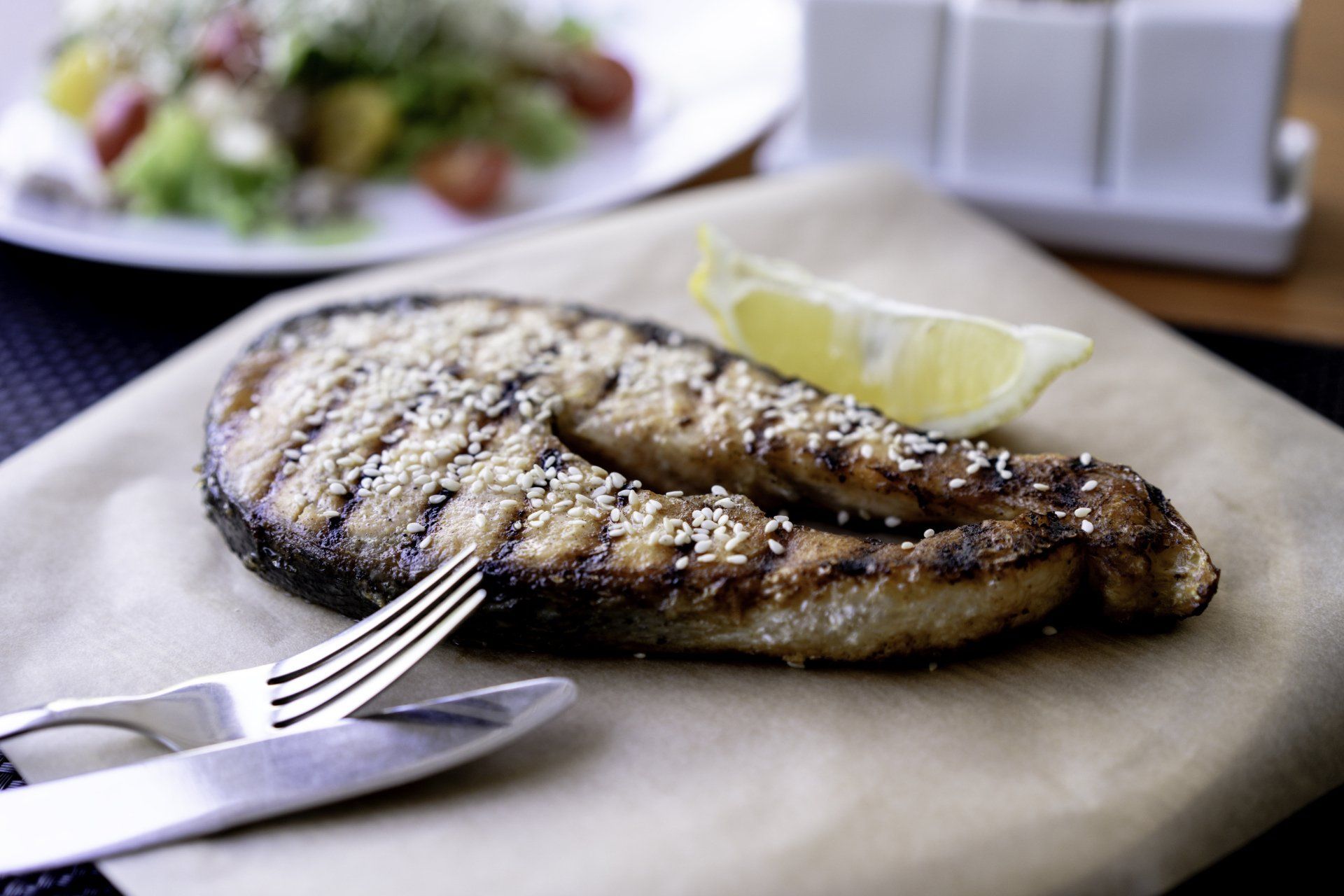 Grilled salmon steak with sesame seeds, lemon wedge, and cutlery on parchment paper; a salad in the background.