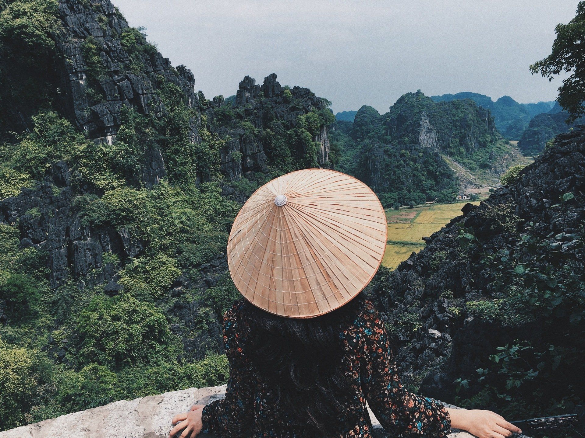 A woman wearing a conical hat is looking out over a mountain range.