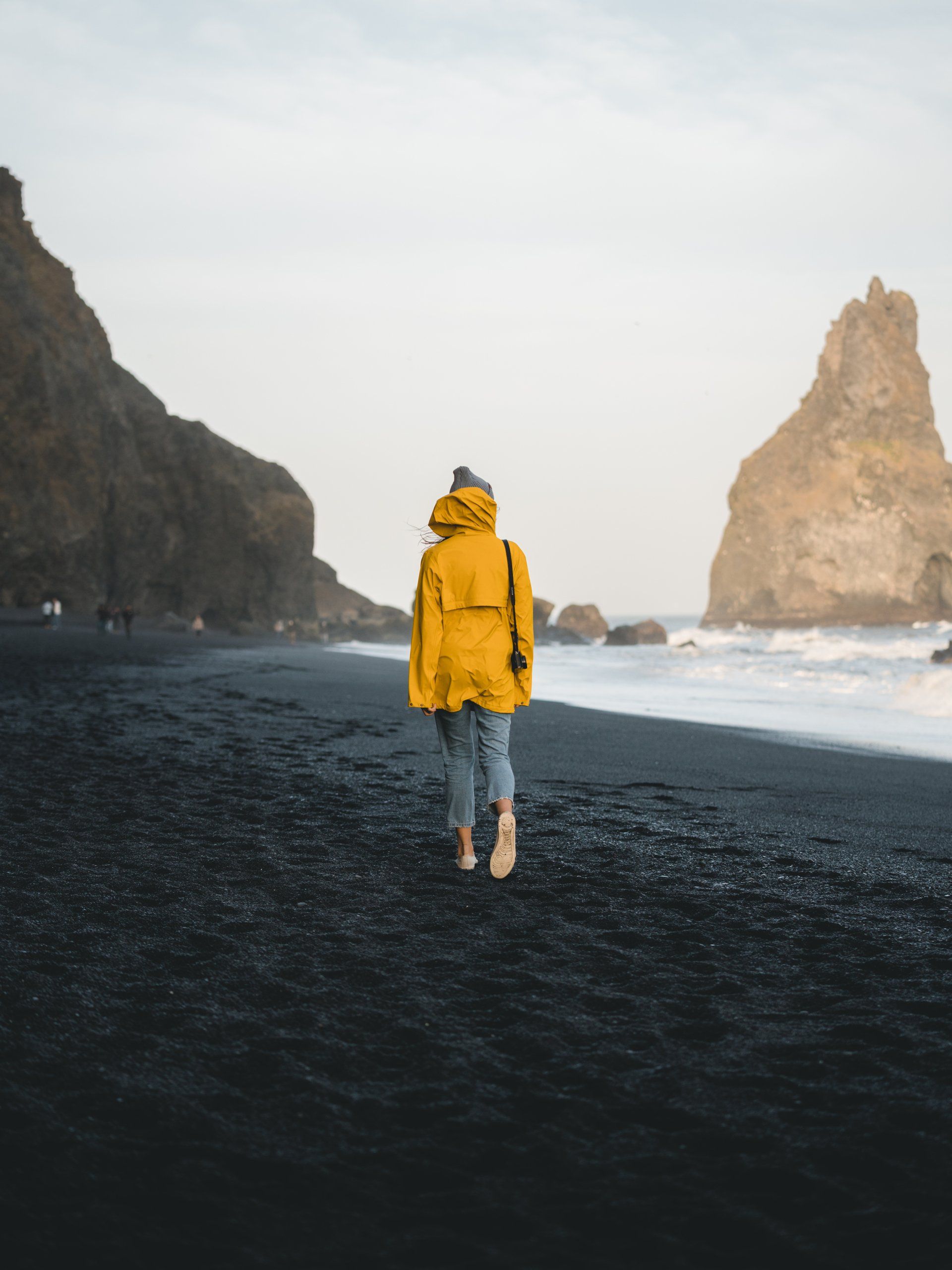 Eine Person in einer gelben Jacke läuft an einem schwarzen Sandstrand entlang.