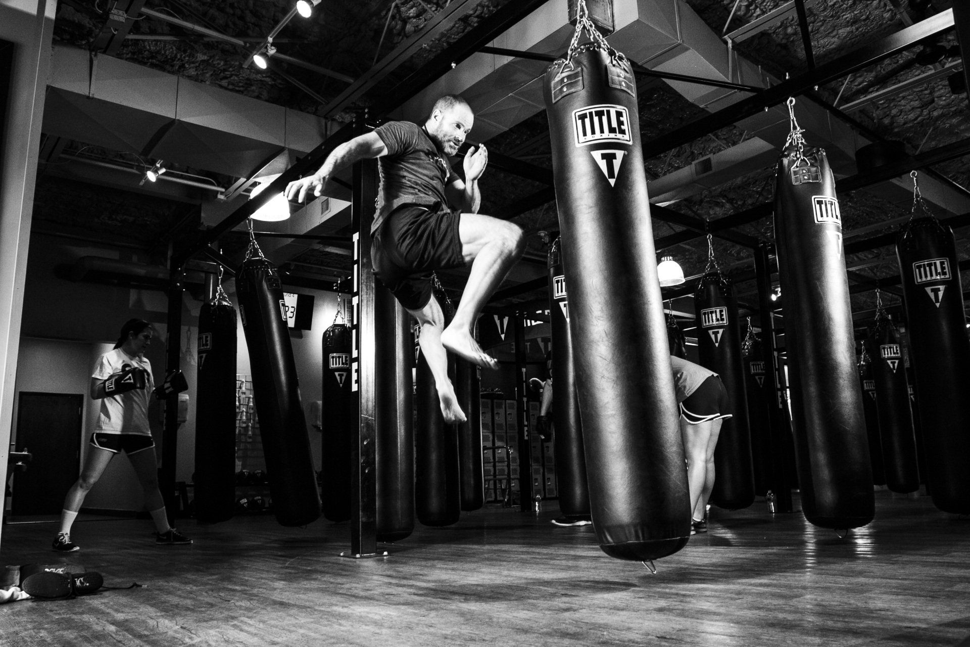 Man jumps to kick heavy bag in a gym, others practice boxing. Black and white.
