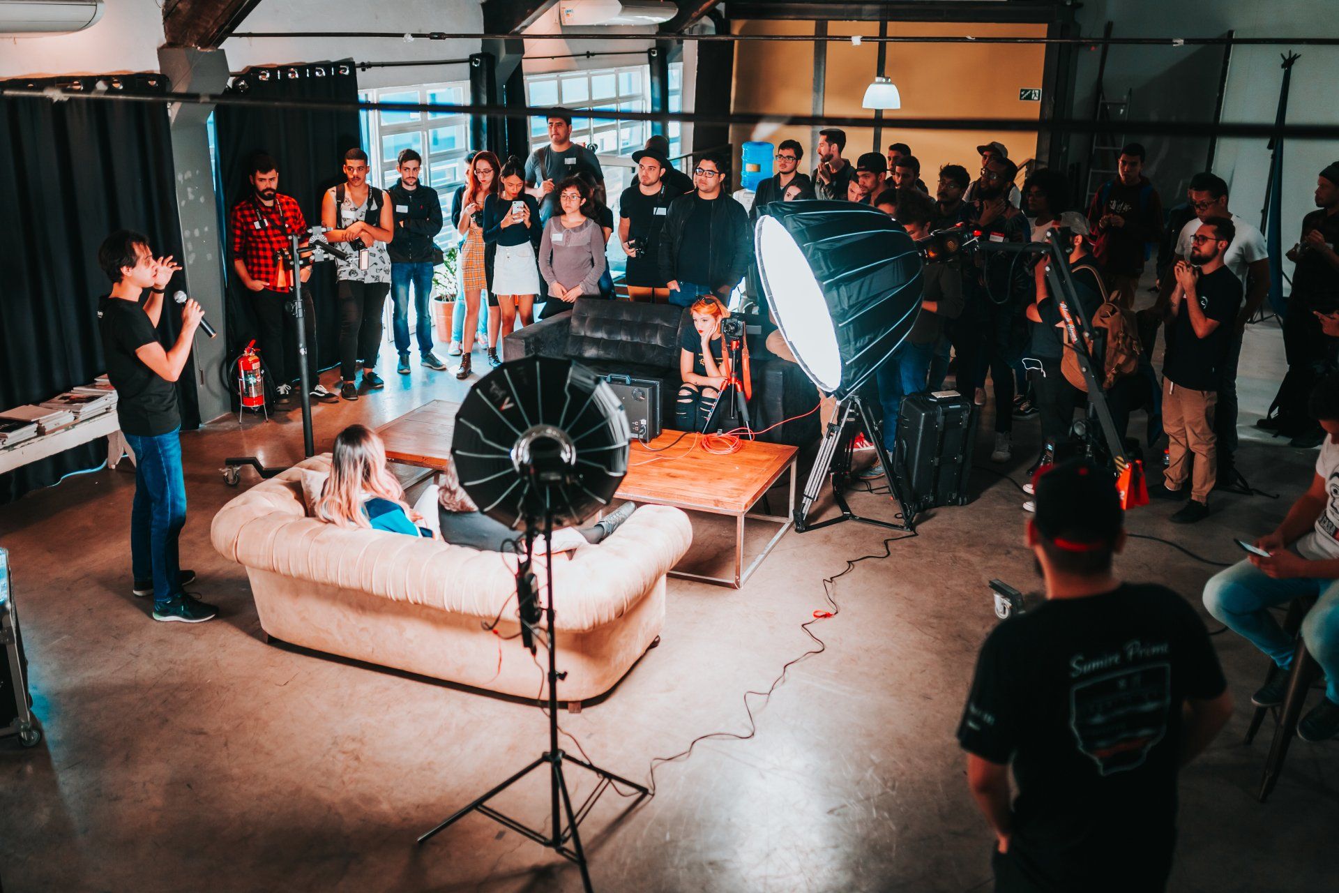 Indoor film set with couch, lights, and crew, actors speaking to the camera and audience.