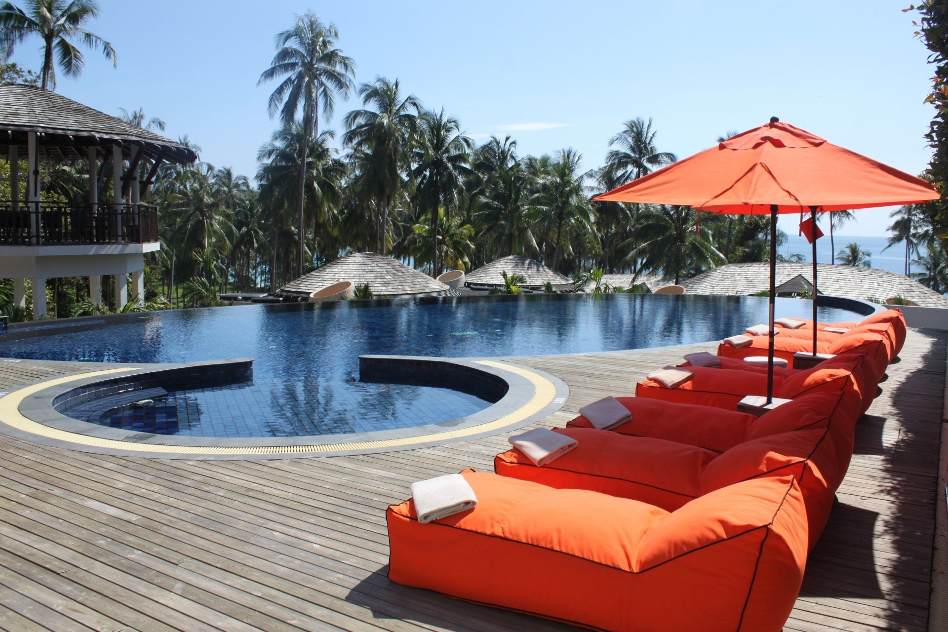 Piscina do resort com espreguiçadeiras e guarda-sóis laranja ao lado de palmeiras e uma lagoa azul.