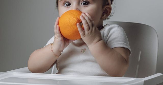 A baby is sitting in a high chair eating an orange.