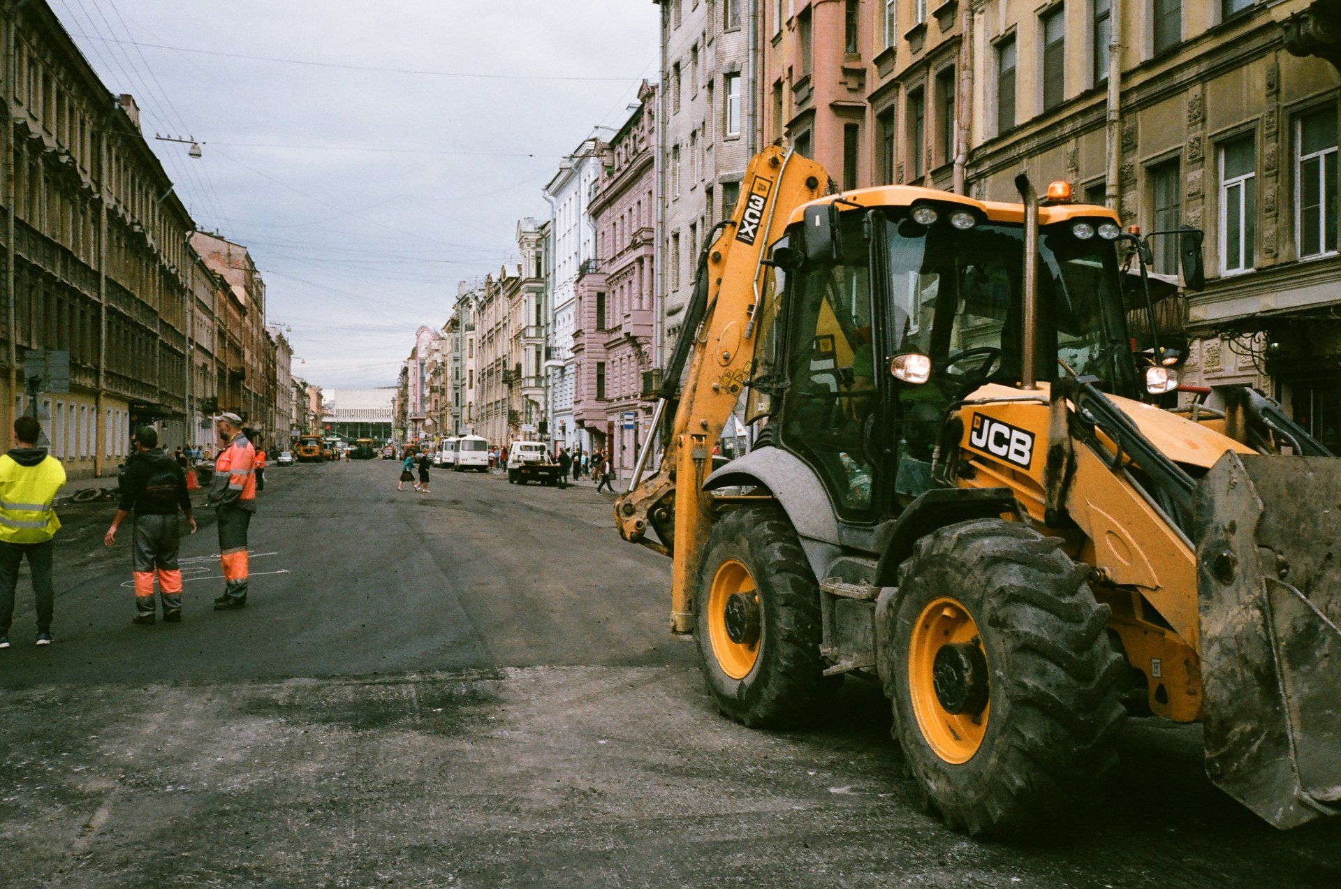 Construction site on a city street; backhoe loader and workers, old colorful buildings in the background.