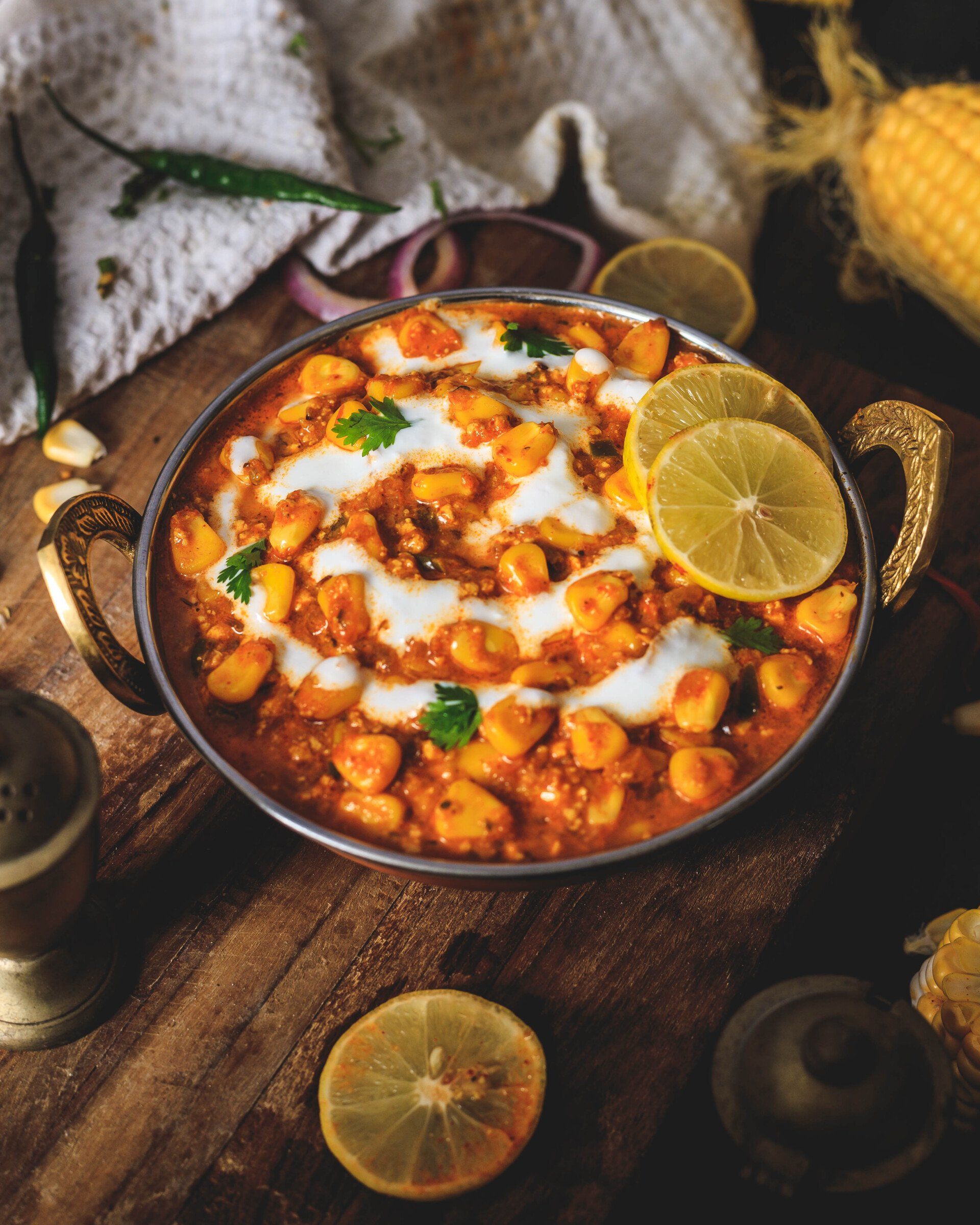 Creamy corn dish in a metal bowl, garnished with lemon slices and cilantro, on a wooden surface.