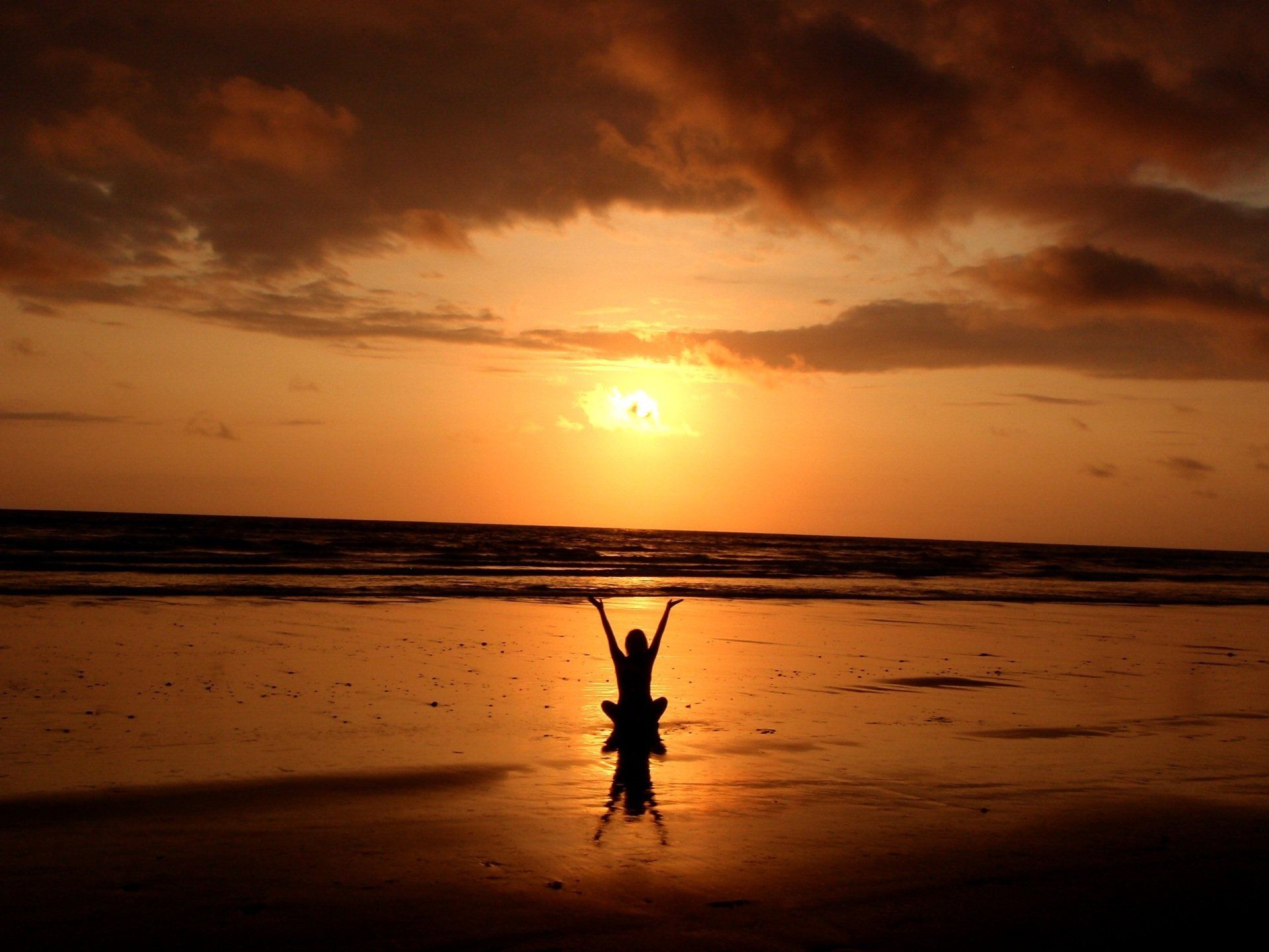 Person on beach with arms raised, silhouetted against golden sunset over ocean.