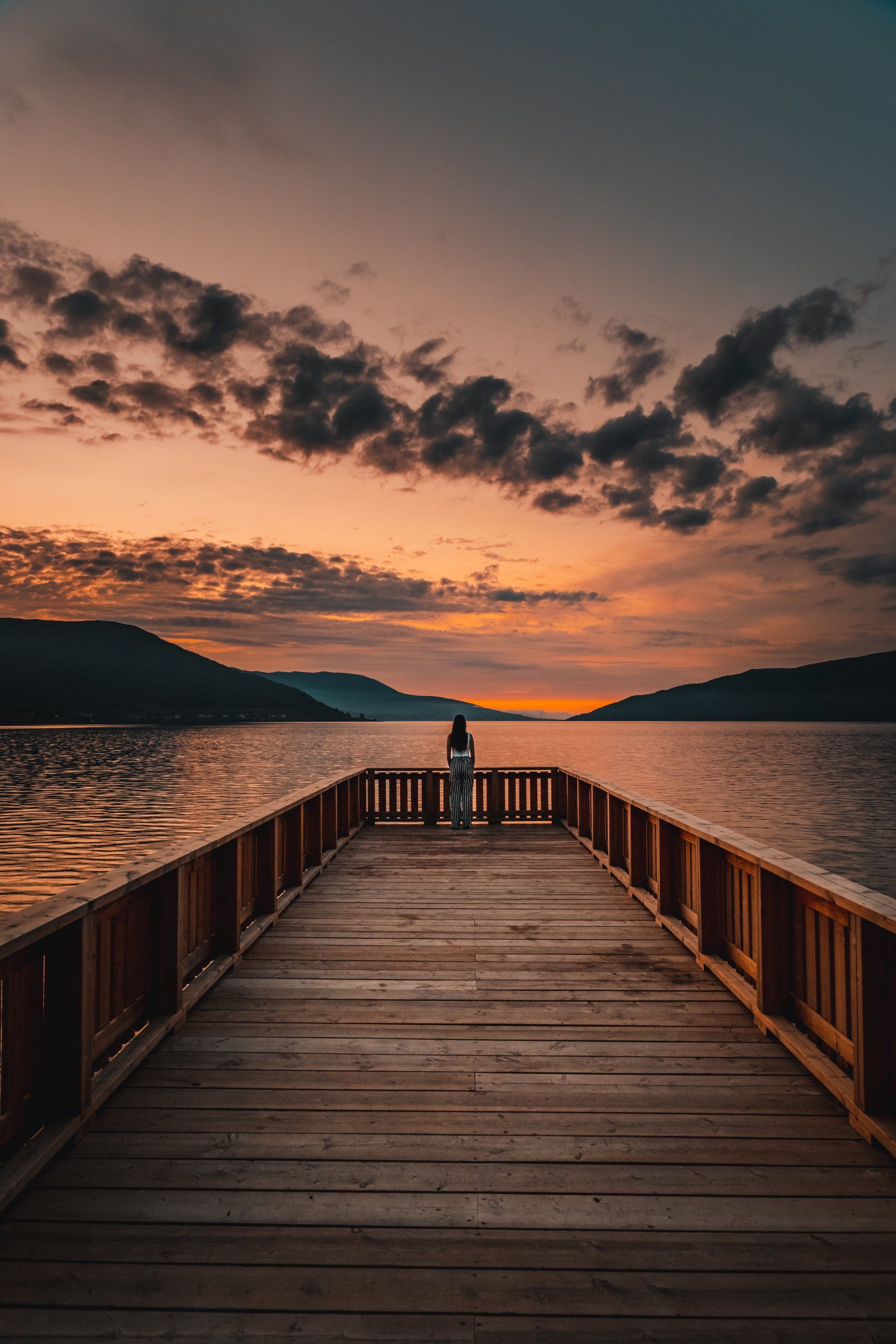 A woman is standing on a wooden pier overlooking a lake at sunset.