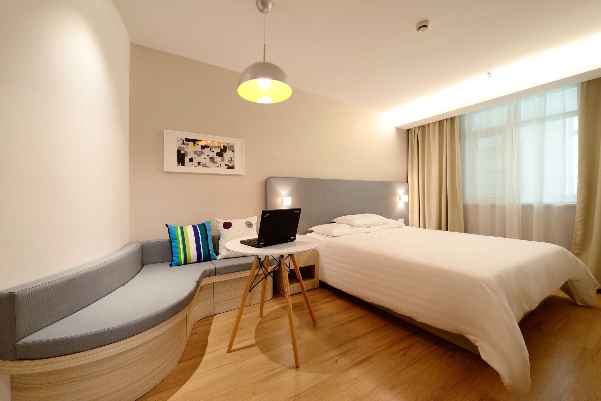 Bedroom with bed, small desk with laptop, built-in curved bench, and large window with curtains.