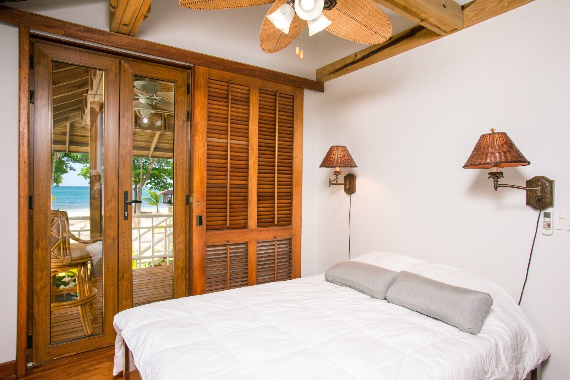 Quarto luminoso com cama branca, vigas de madeira, portas de armário venezianas e portas de vidro para uma varanda.