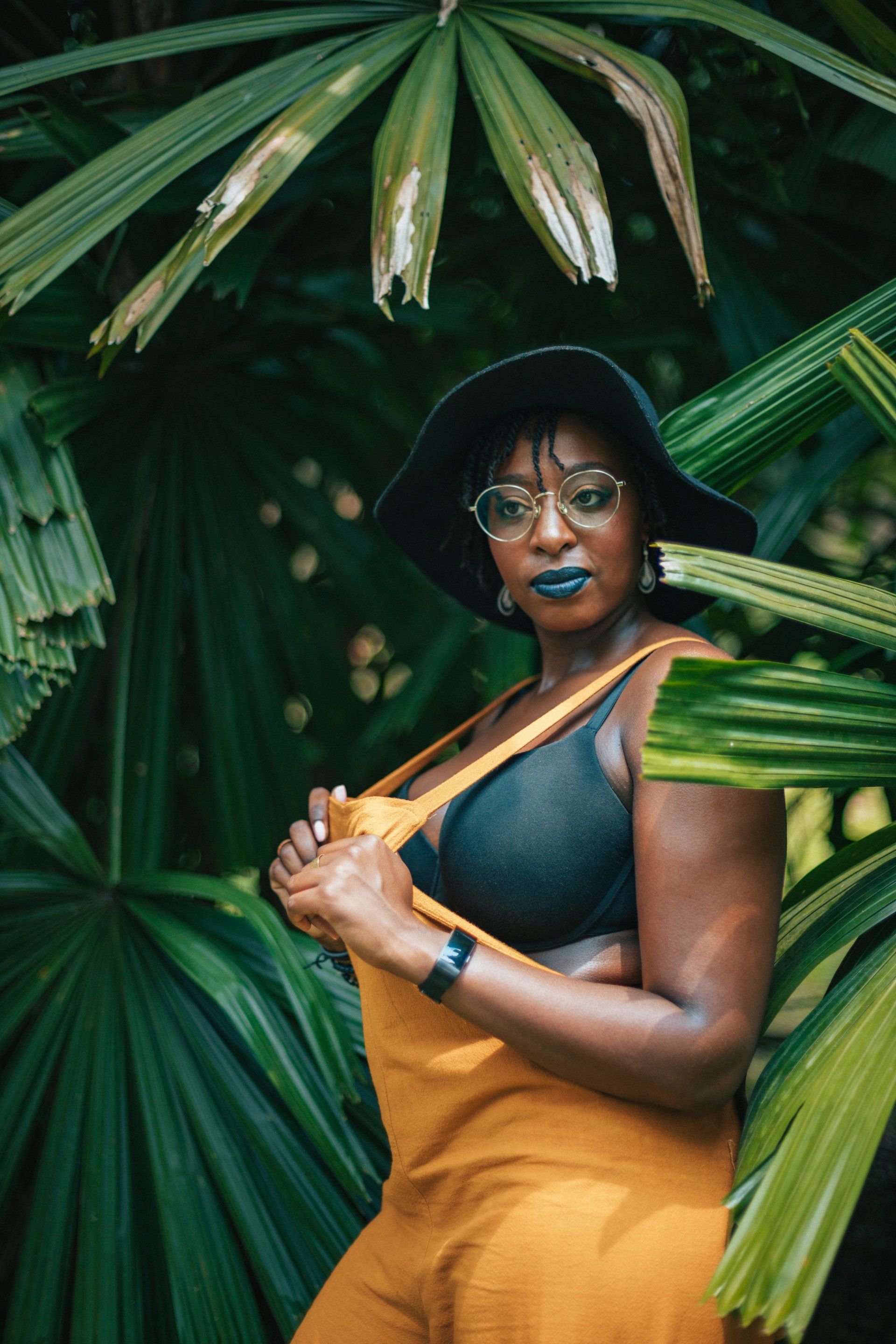 Woman in yellow overalls and black bra, wearing a hat and glasses, posing among green foliage.