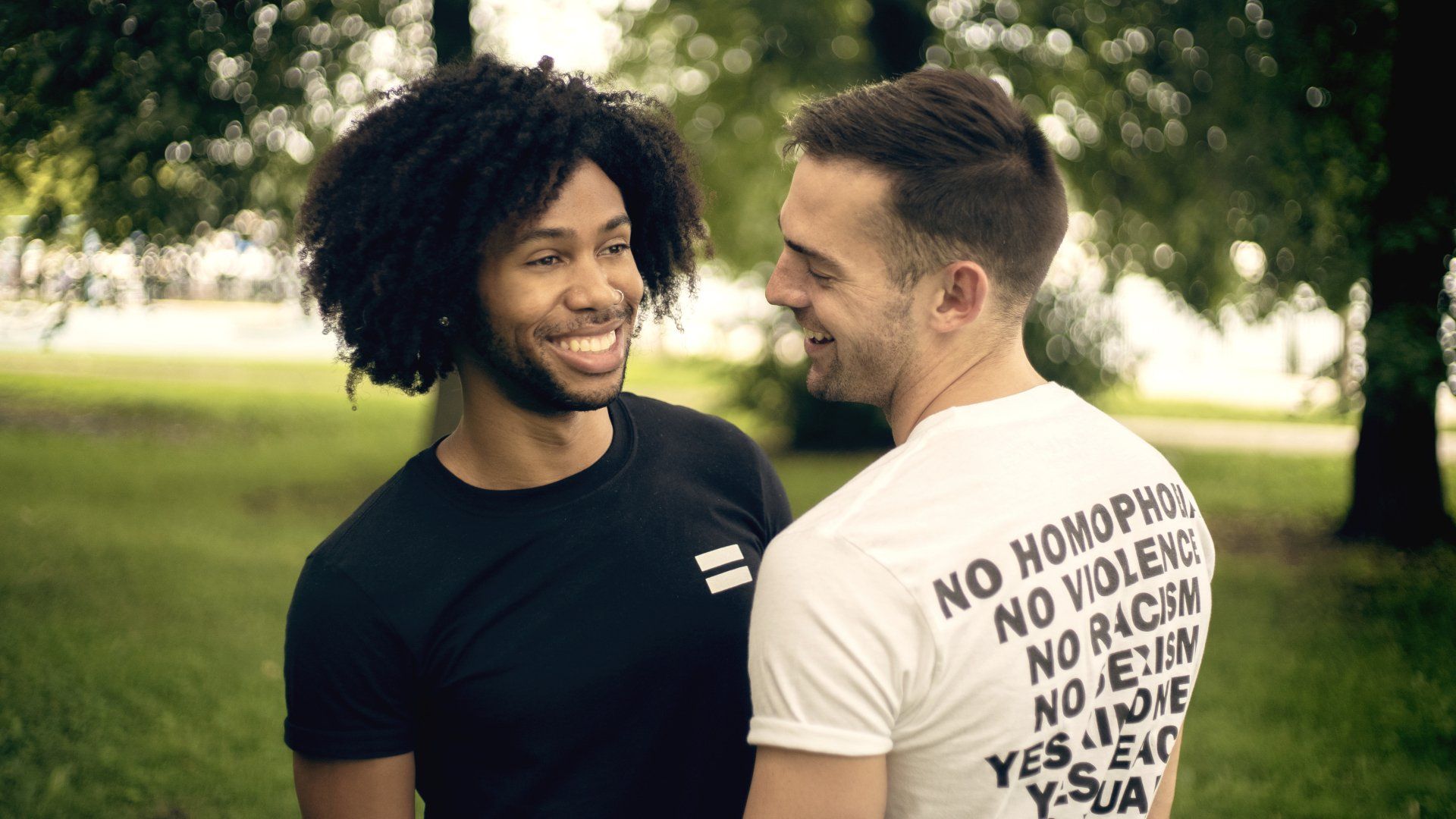 Two men smiling at each other in a park. One wears a black shirt with an equal sign, the other a white shirt with text.